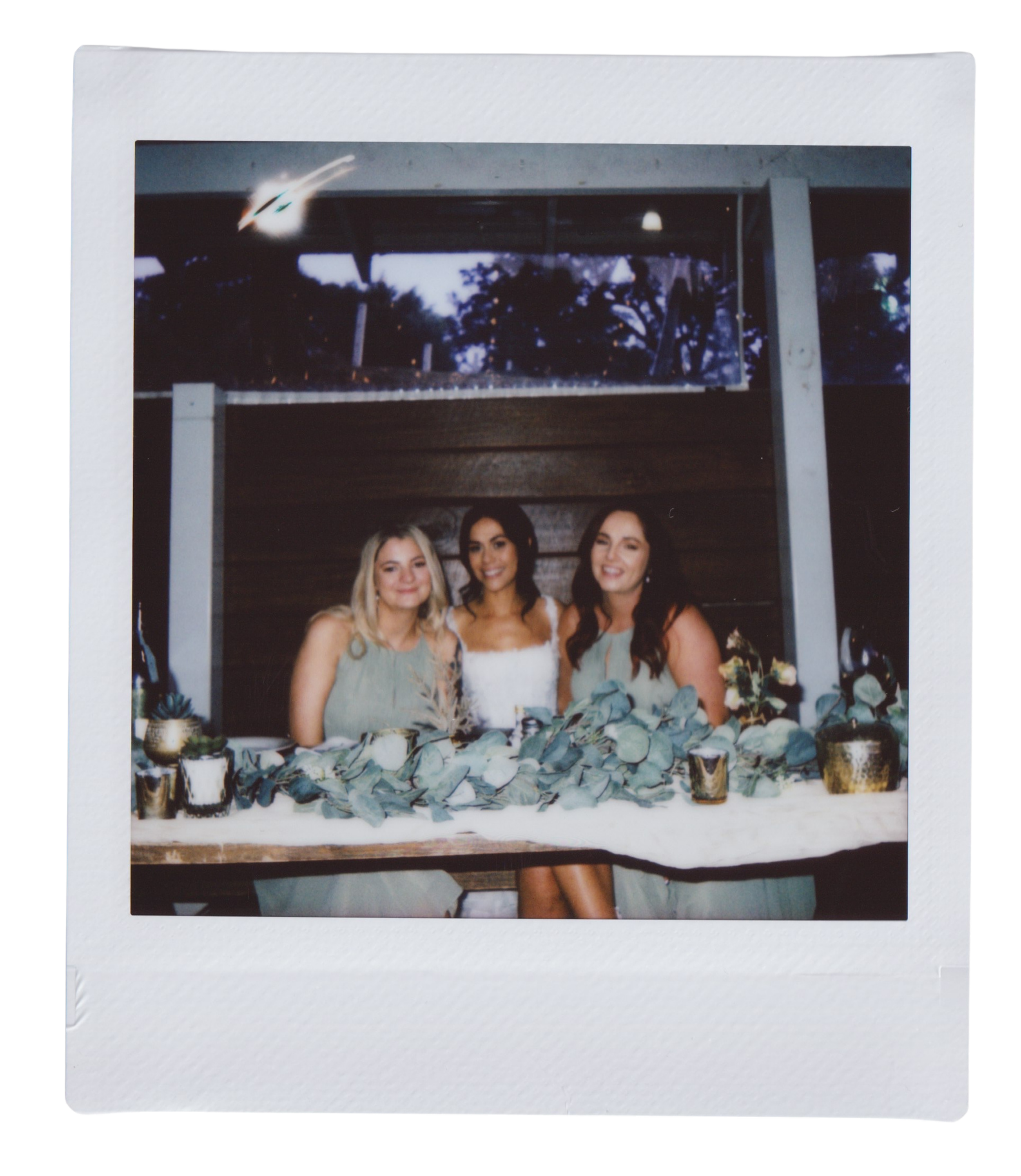 Photograph of three women standing behind a floral table setting, with a white box frame around the photo and trees visible in the background.