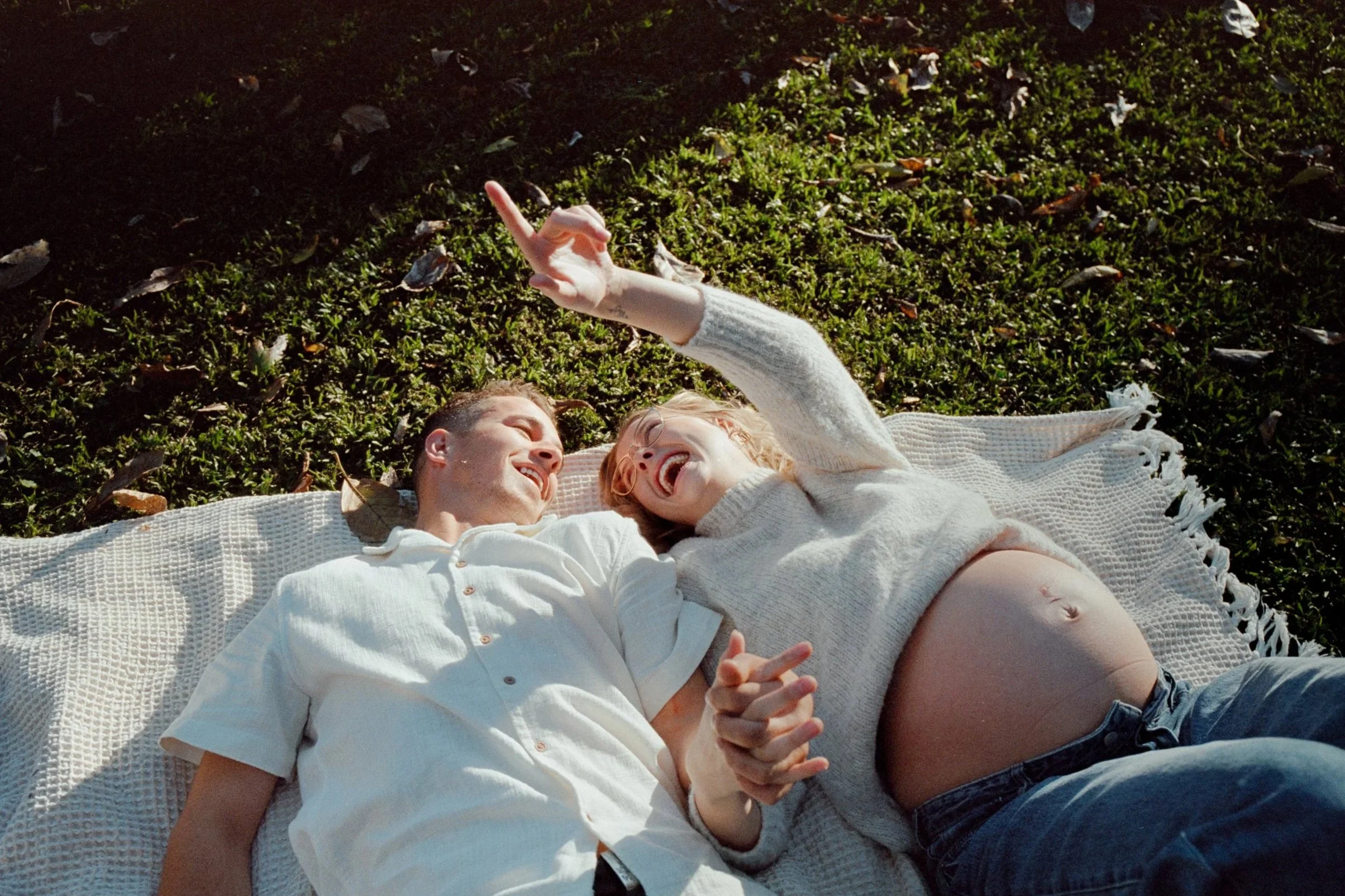 Two people, a man and a woman, lying on a blanket outdoors on green grass, smiling and laughing, with the woman making a peace sign, during daytime.