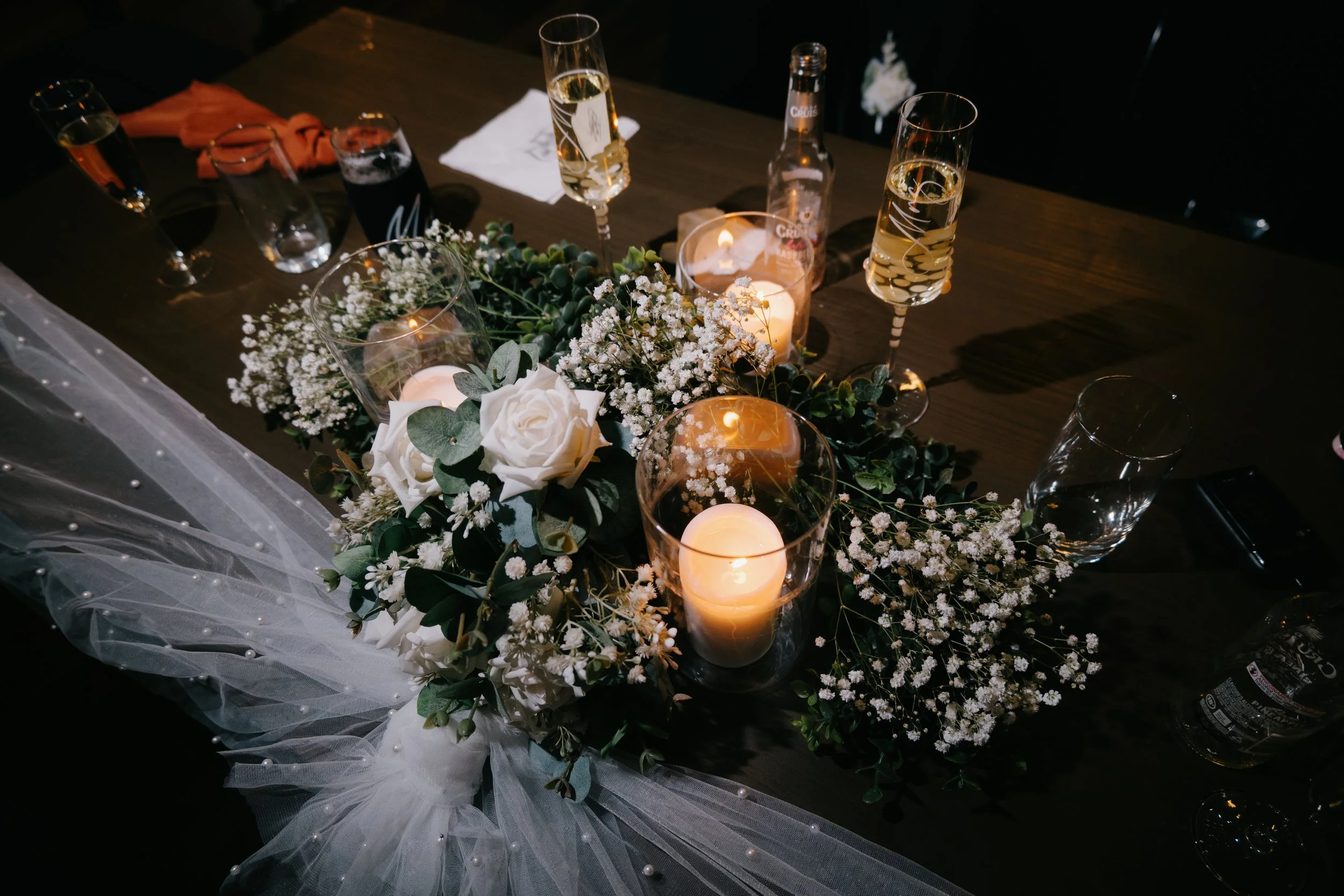 Decorative table centerpiece with white roses, baby's breath, greenery, candles, and champagne glasses on a dark table at an event or celebration.