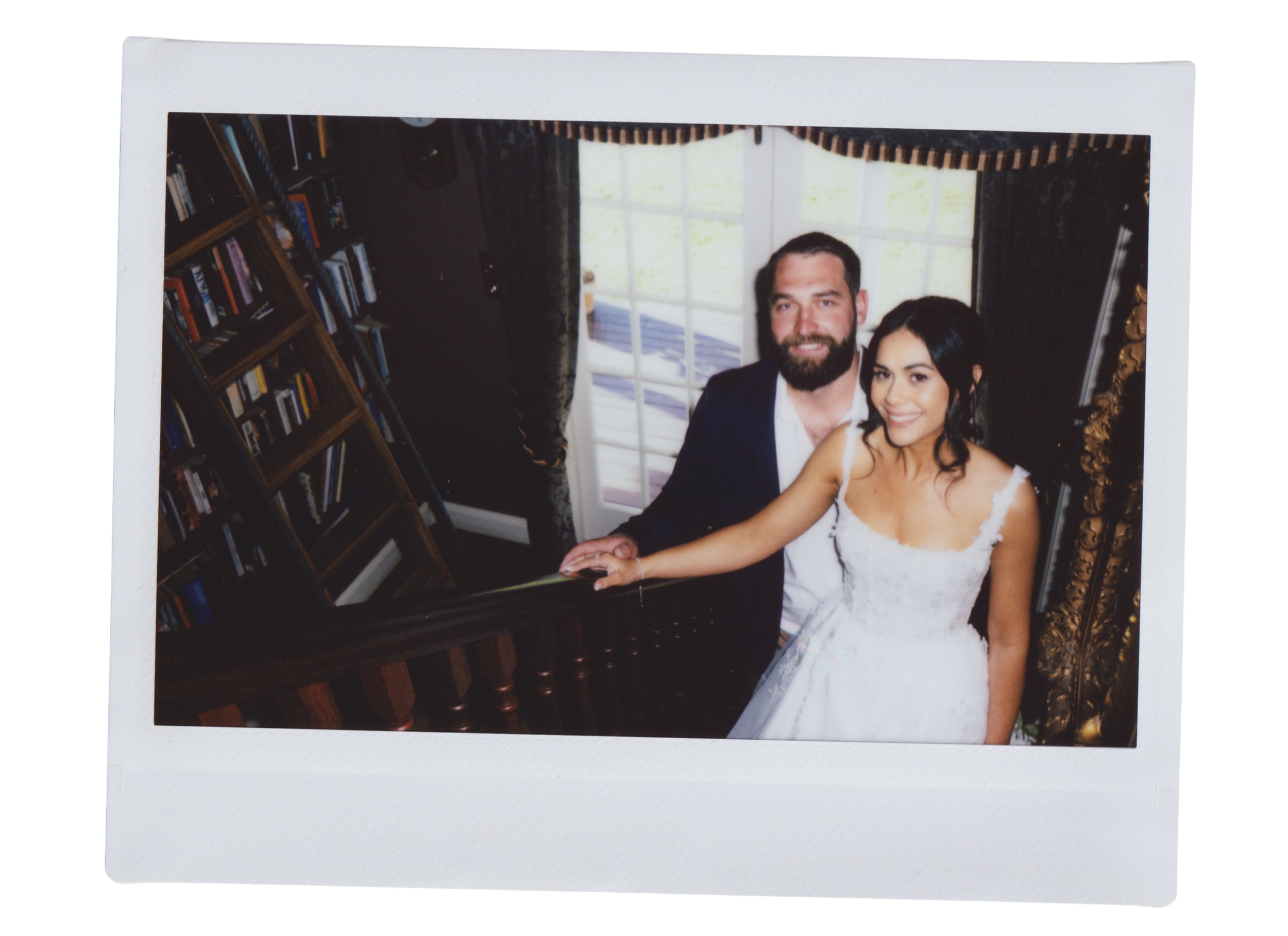 A printed photograph of a smiling couple dressed in formal attire, sitting at a table in a room with bookshelves, a large window, dark curtains, and ornate gold-colored decorations.