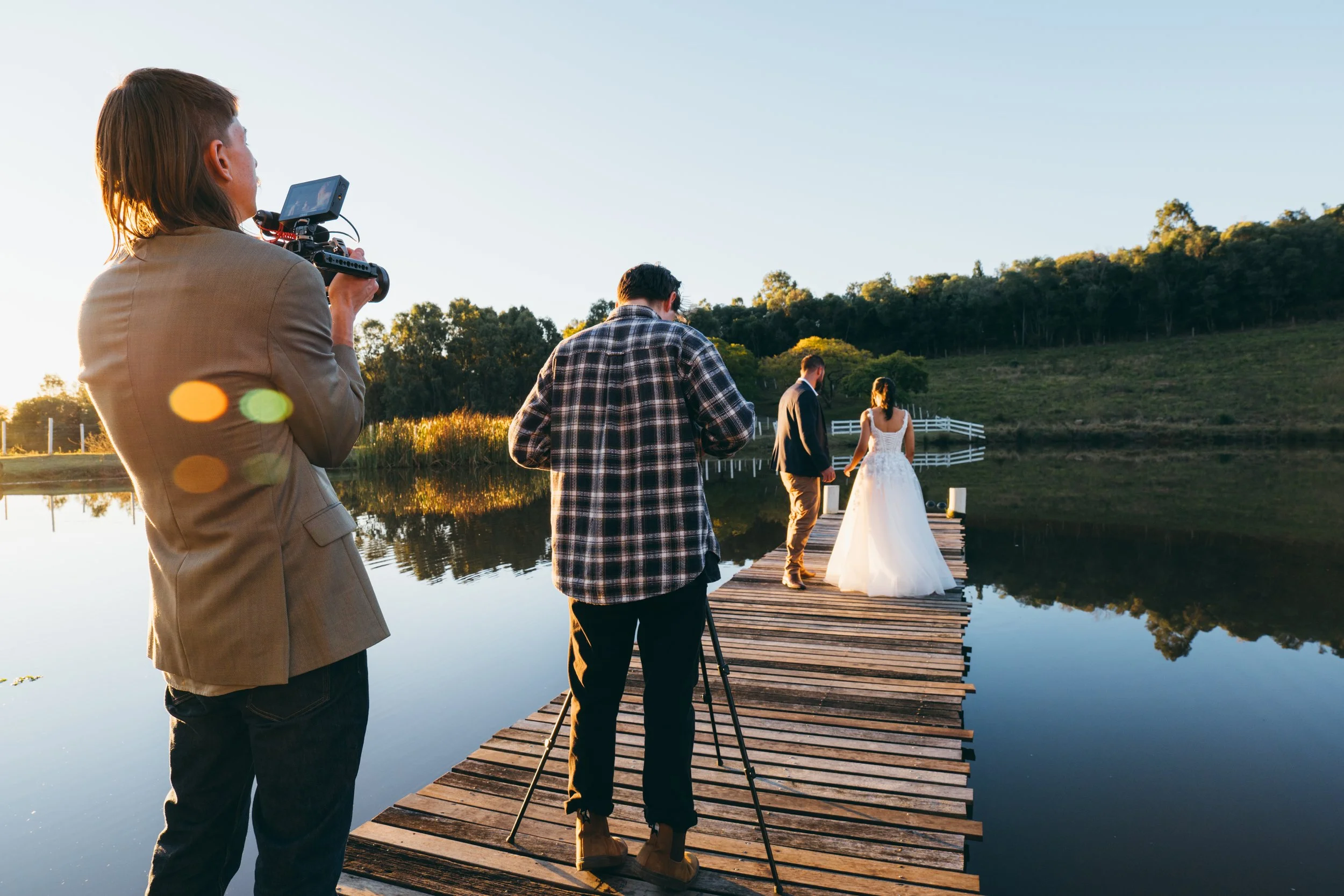 Photographers capturing a wedding couple on a wooden dock by a lake at sunset.