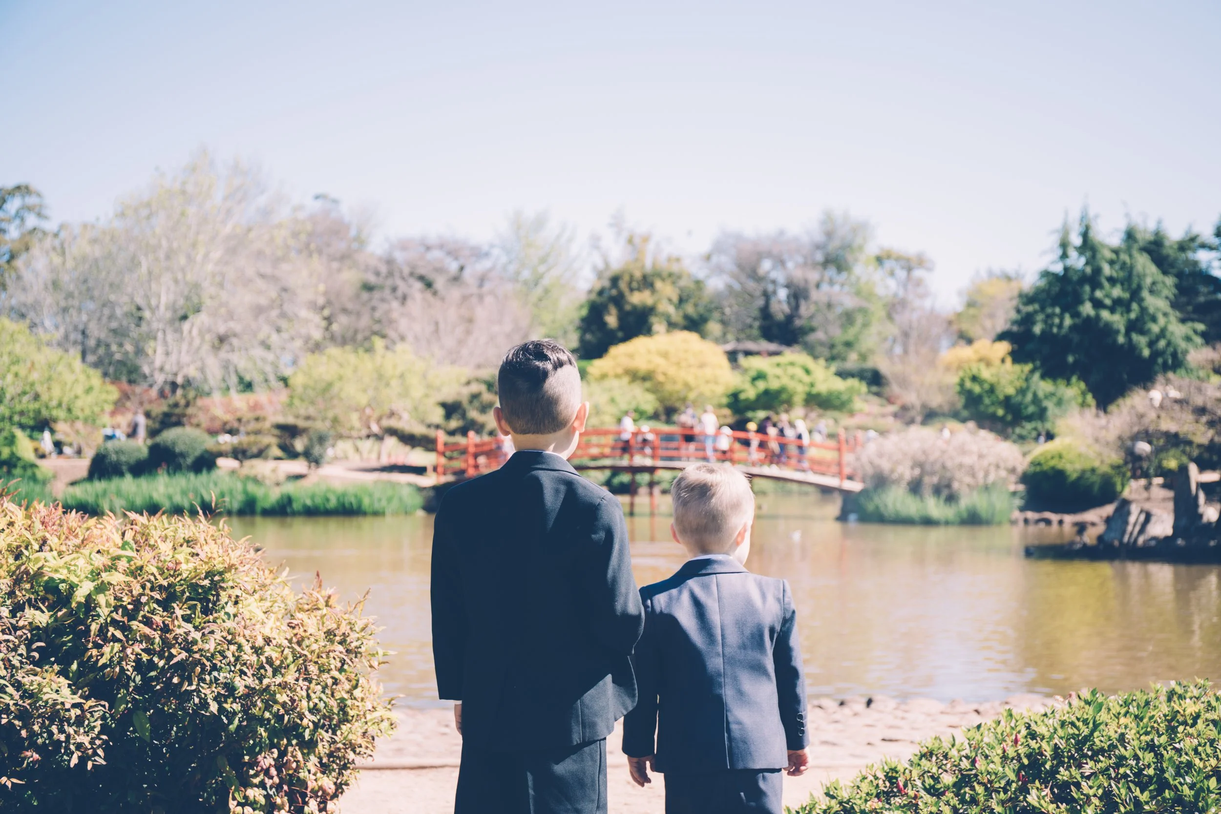 Two boys dressed in suits stand by a pond in a park with trees and a small red bridge in the background.