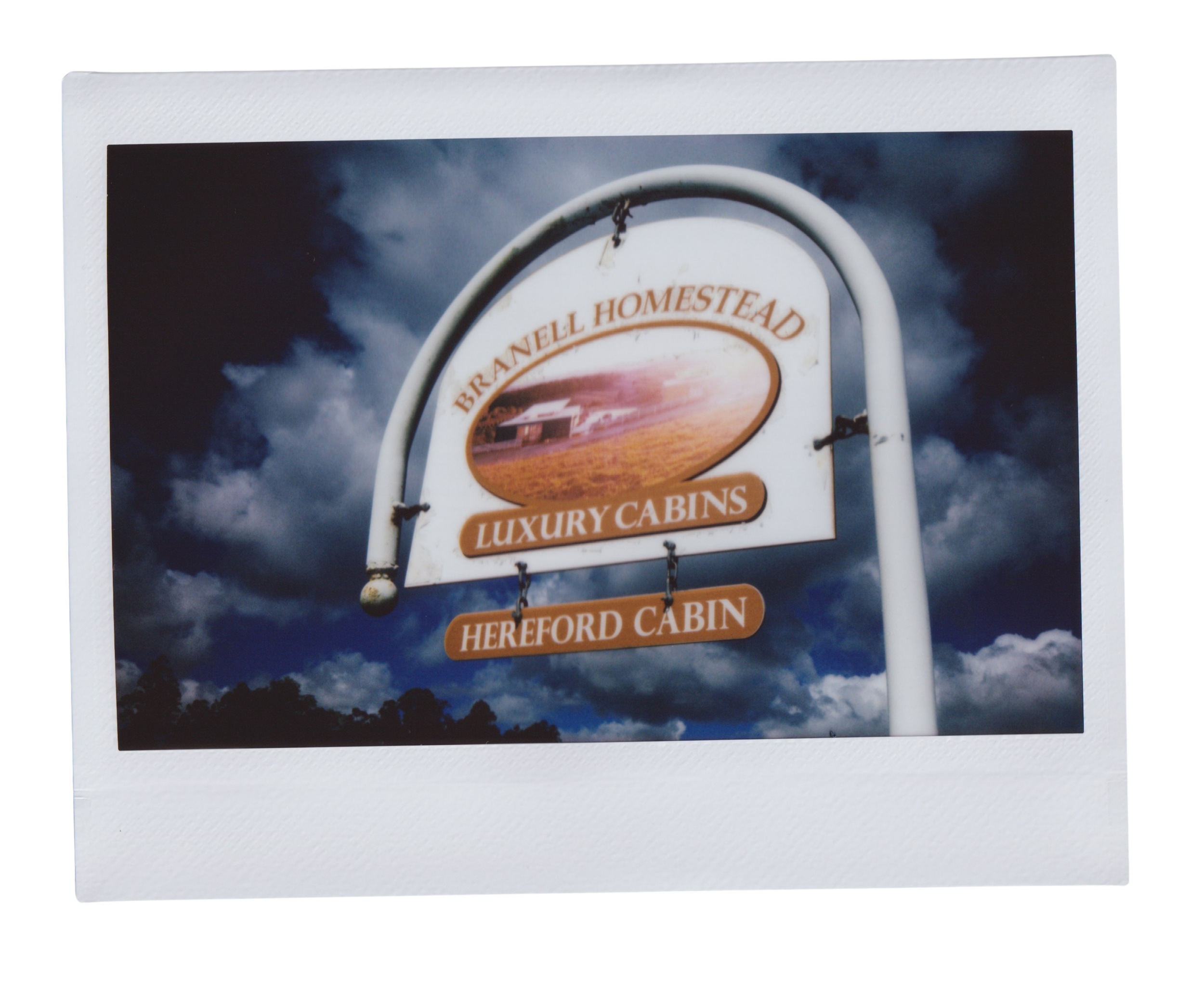 Signboard for Brannel Homestead Luxury Cabins against a cloudy sky with trees below.