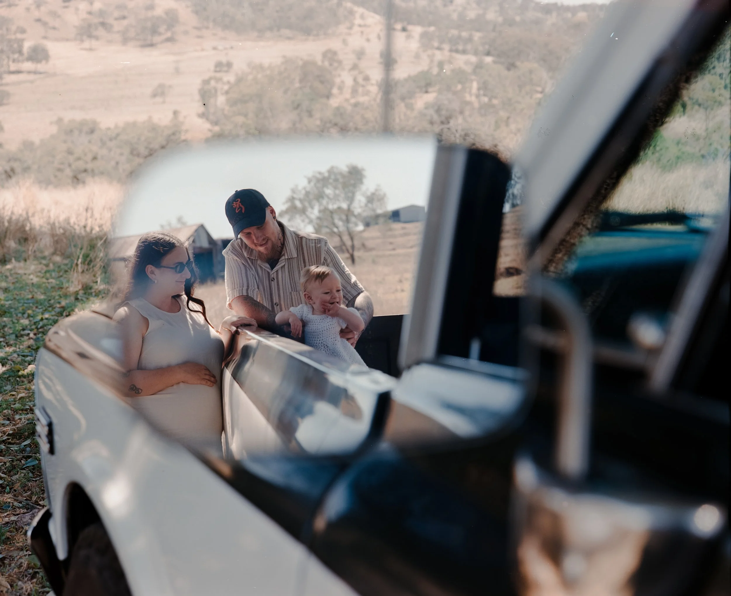 A family of three standing outside near a white vintage car, with a scenic rural landscape and a lake in the background, as seen through the car's side mirror.