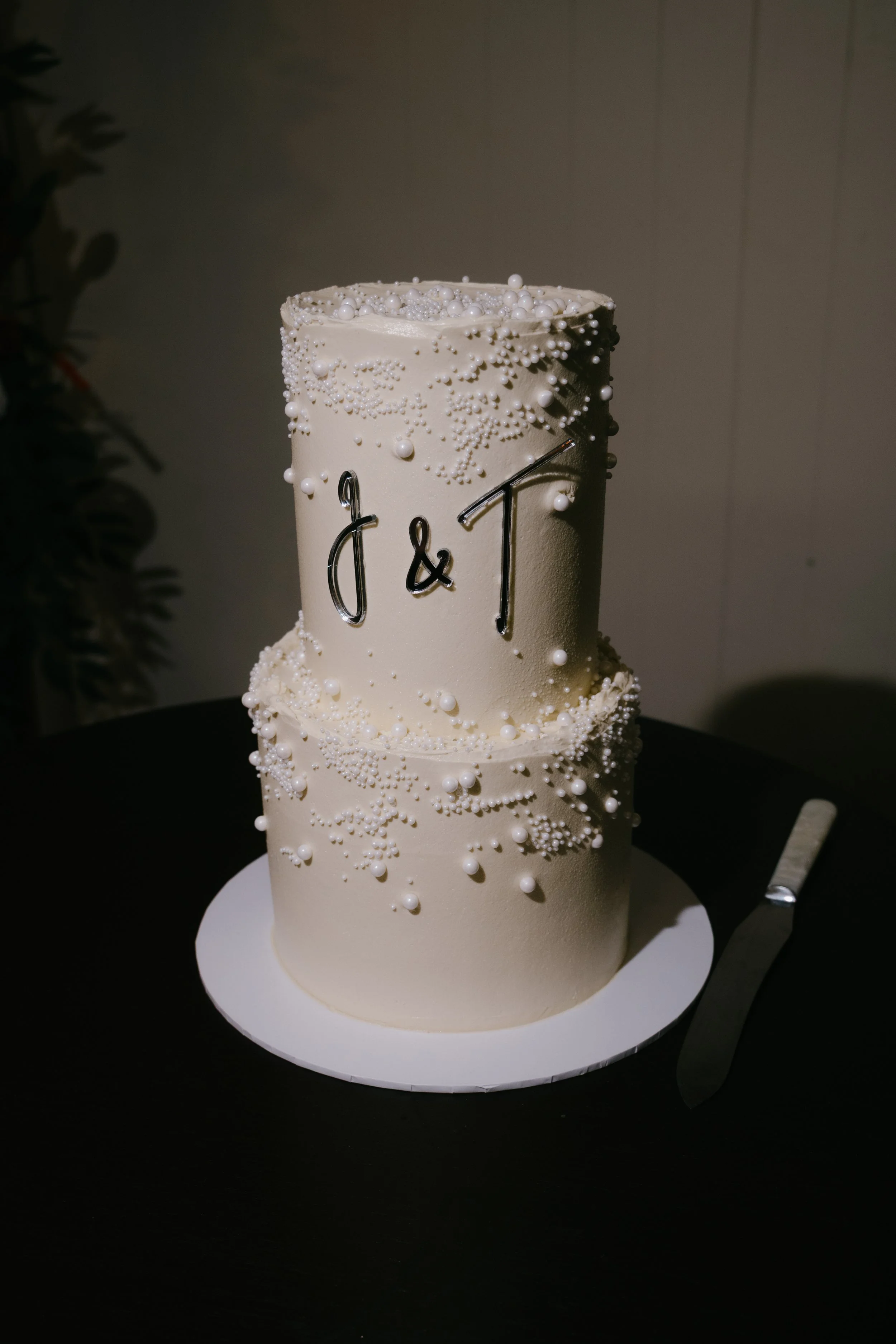 A tall, white, cylindrical cake decorated with small white pearls and beads, featuring the initials 'J & T' in black on the front, placed on a white cake board, themed for a celebration.