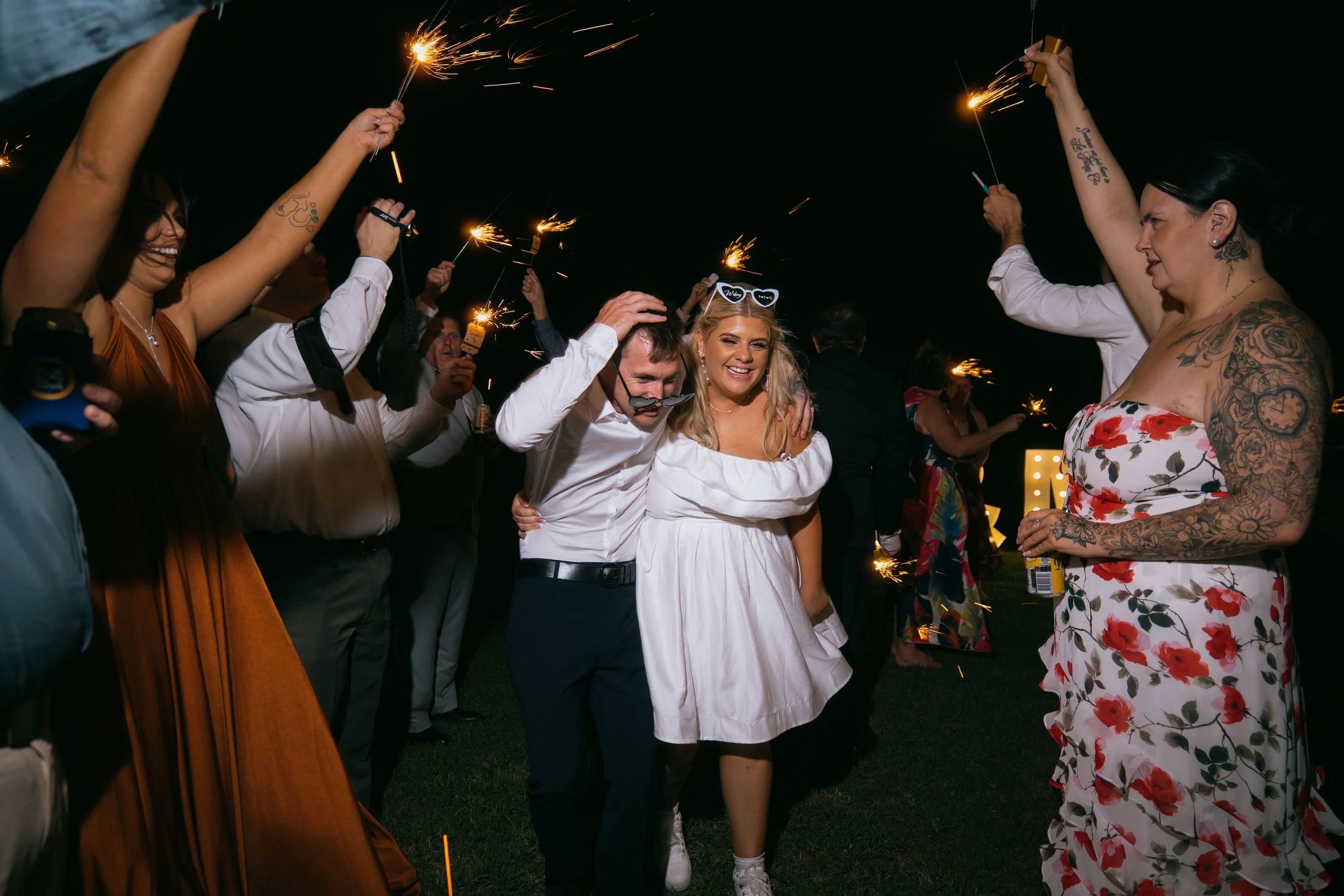 Group of people celebrating at night with sparklers, some smiling and embracing, dressed in casual and semi-formal attire.