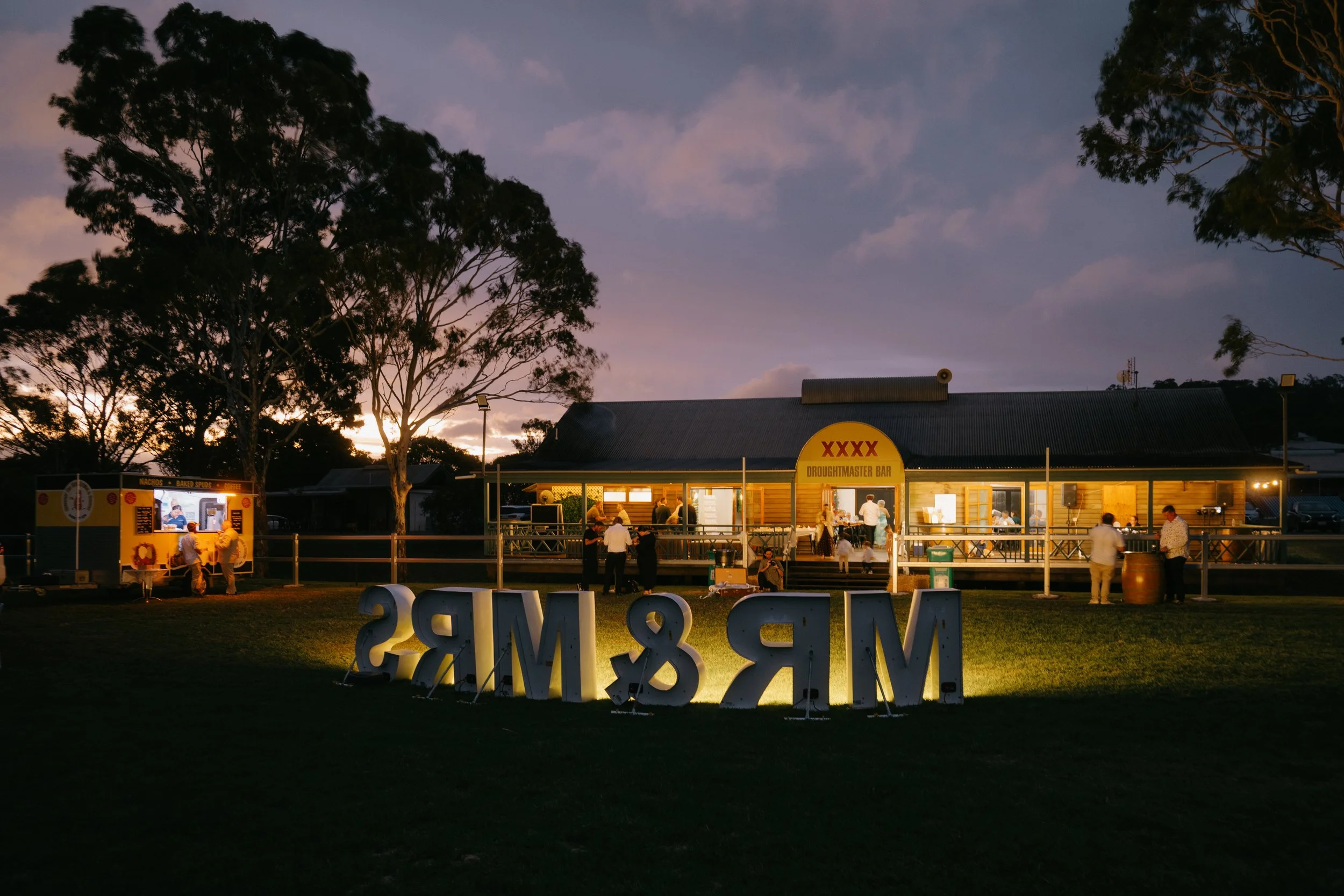 People gathered outside a building with a sign that reads 'XXXX Droughtmaster Bar' during dusk, with trees and a cloudy sky in the background, and large white letters spelling 'MS & MS' on the grass in the foreground.