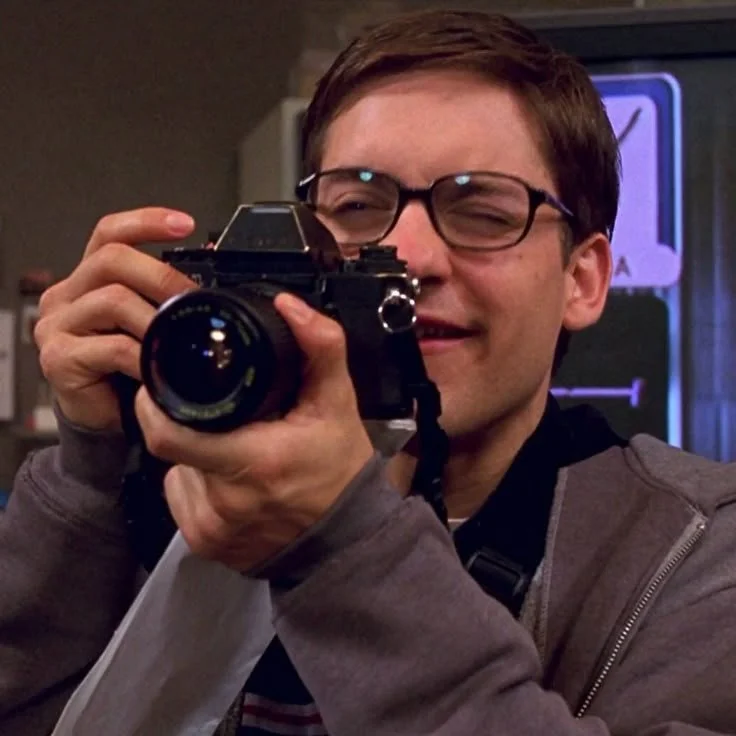 A young man with glasses and a gray jacket taking a photo with a vintage camera indoors.