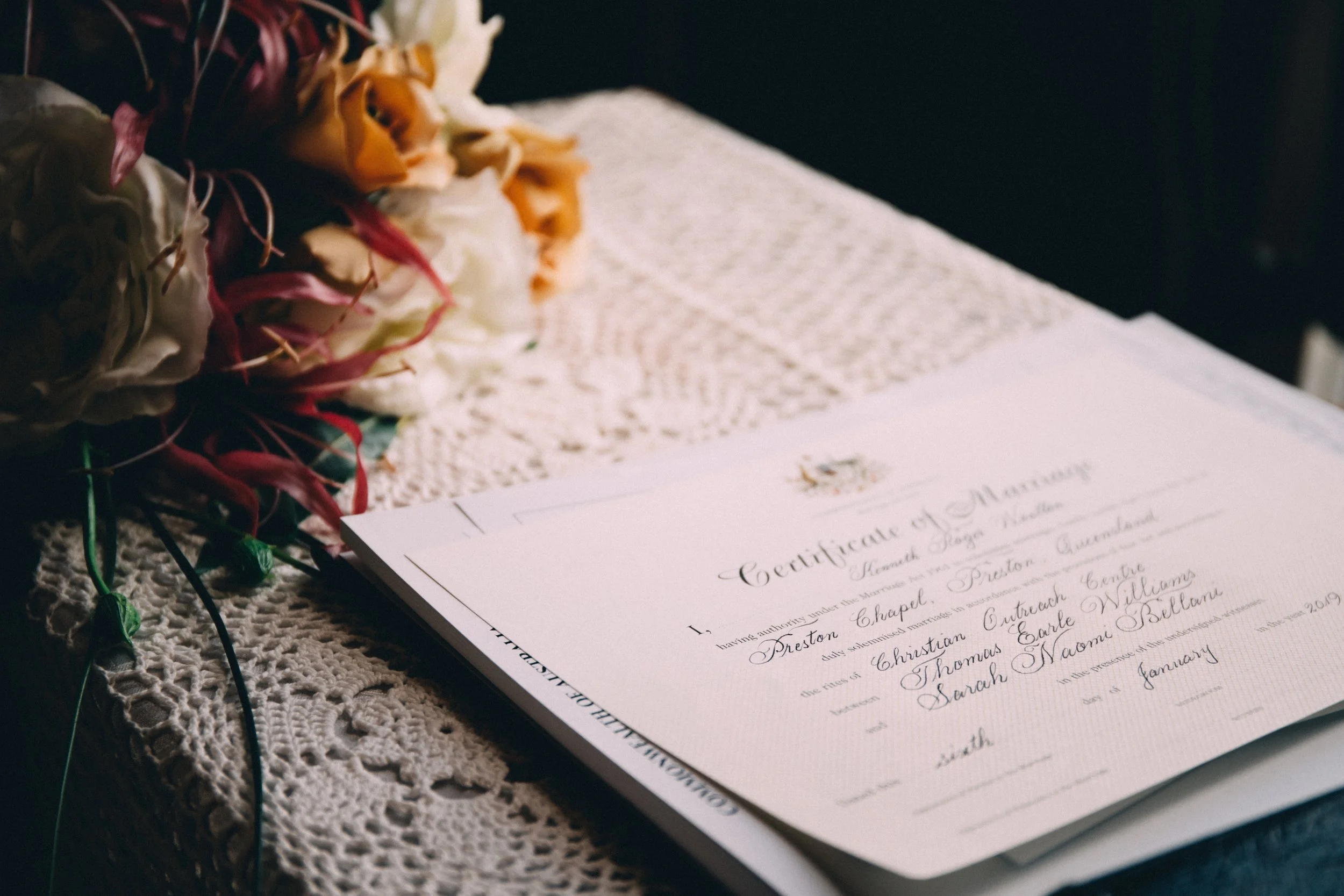 Close-up photo of a wedding certificate and a bouquet of flowers on a lace tablecloth.