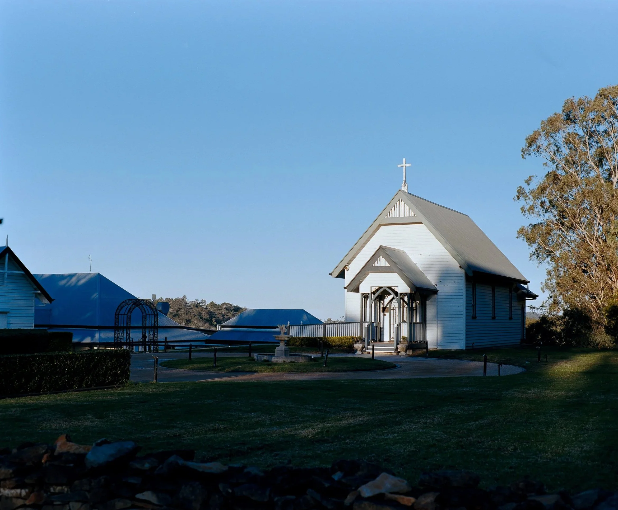 A white church with a cross on top, surrounded by trees and a well-kept lawn, under a clear blue sky.