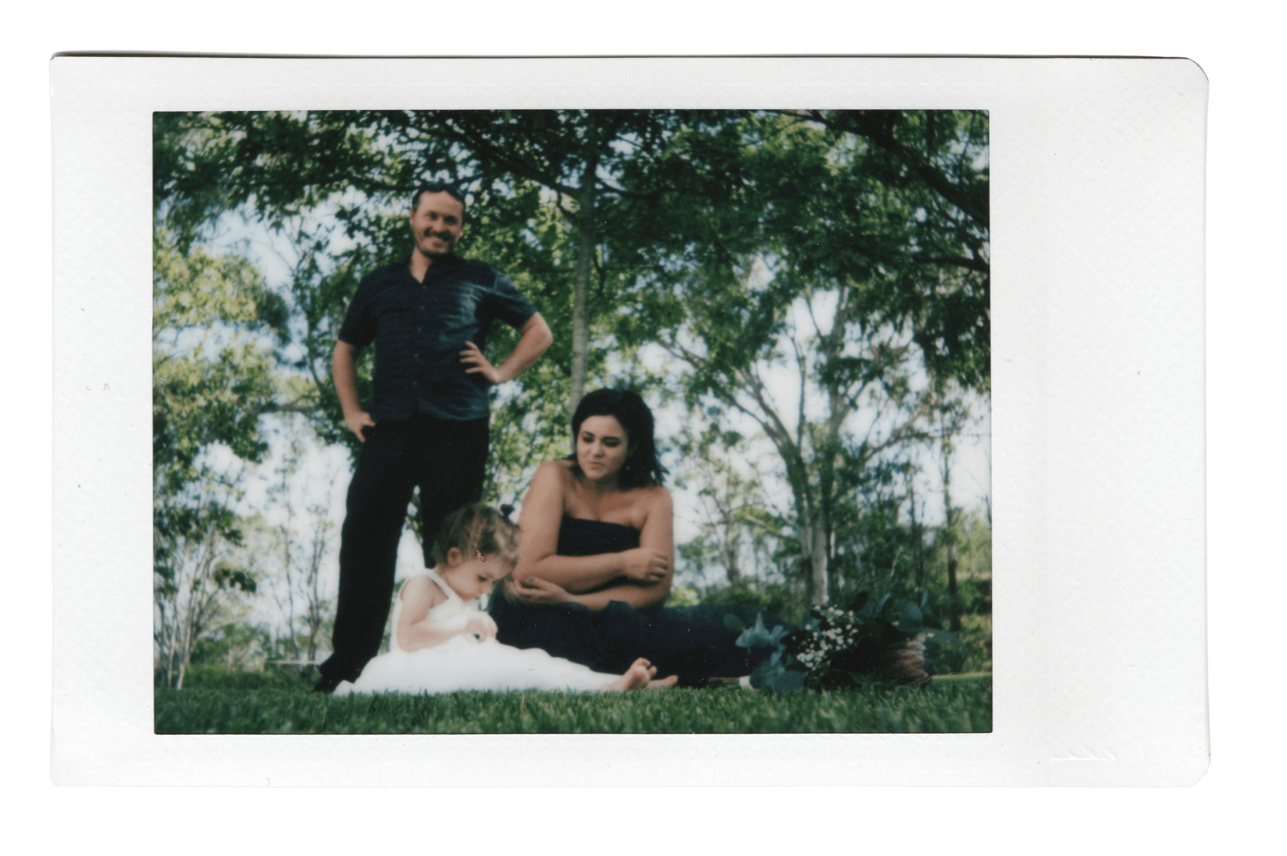 A printed photo of a group of people outdoors in a park. An adult man is standing with one hand on his hip, smiling. A woman is sitting on the grass holding two children, one on each side. The scene is surrounded by trees and green foliage.