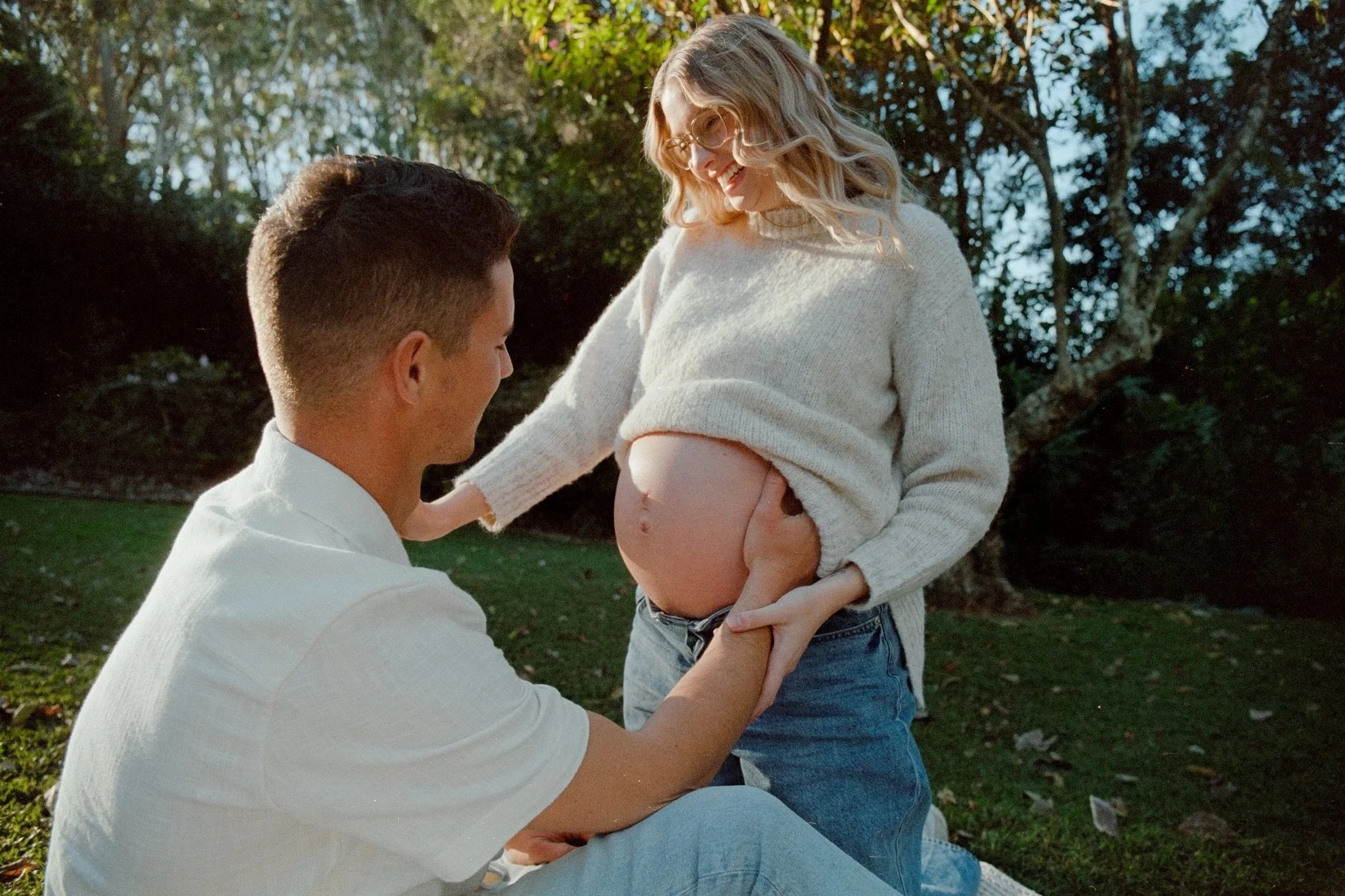 A man kneeling in front of a pregnant woman outdoors, holding her belly, with a backdrop of trees and sunlight filtering through the leaves.