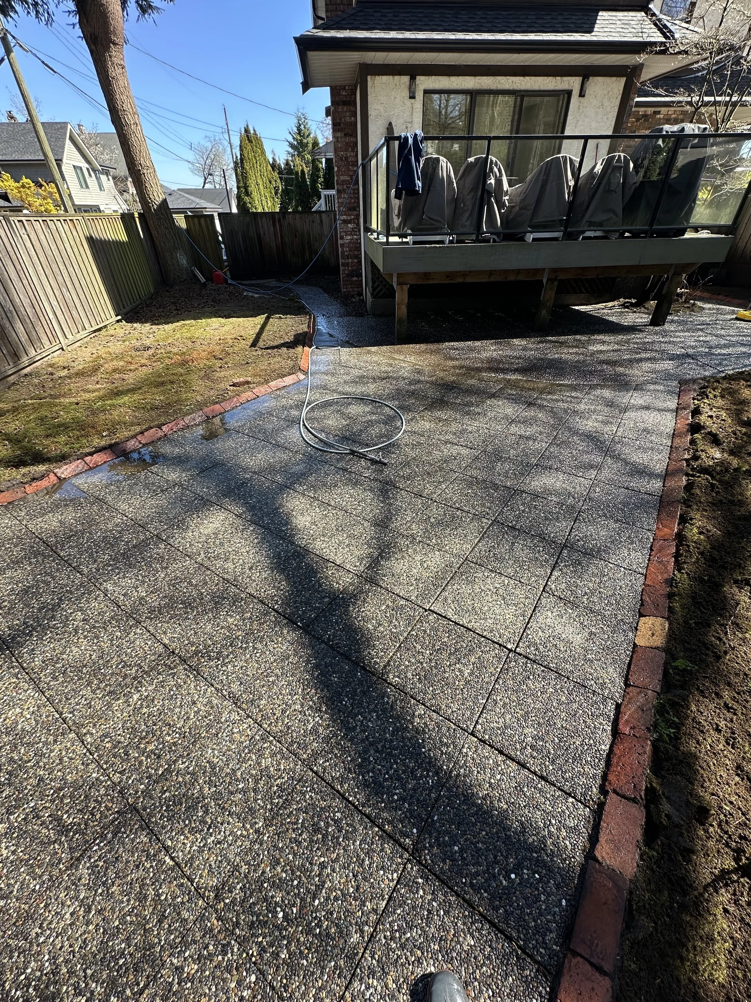 Backyard patio with wet paving stones, a lawn, a wooden fence, a house with a balcony with covered outdoor furniture, and a tree with a water hose on the ground.