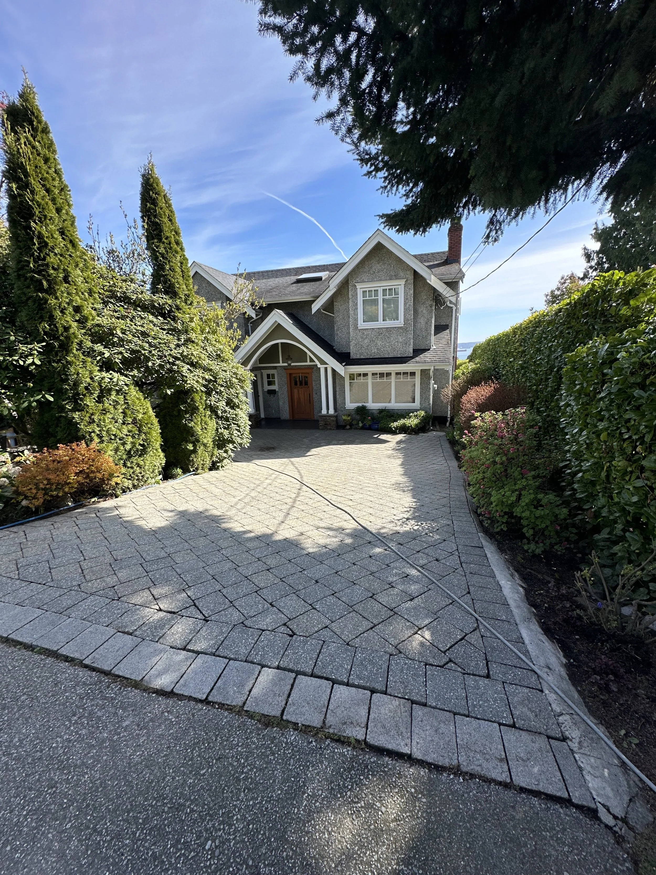 A paved driveway leading to a house with a gray exterior, surrounded by green bushes and trees, with a blue sky overhead.