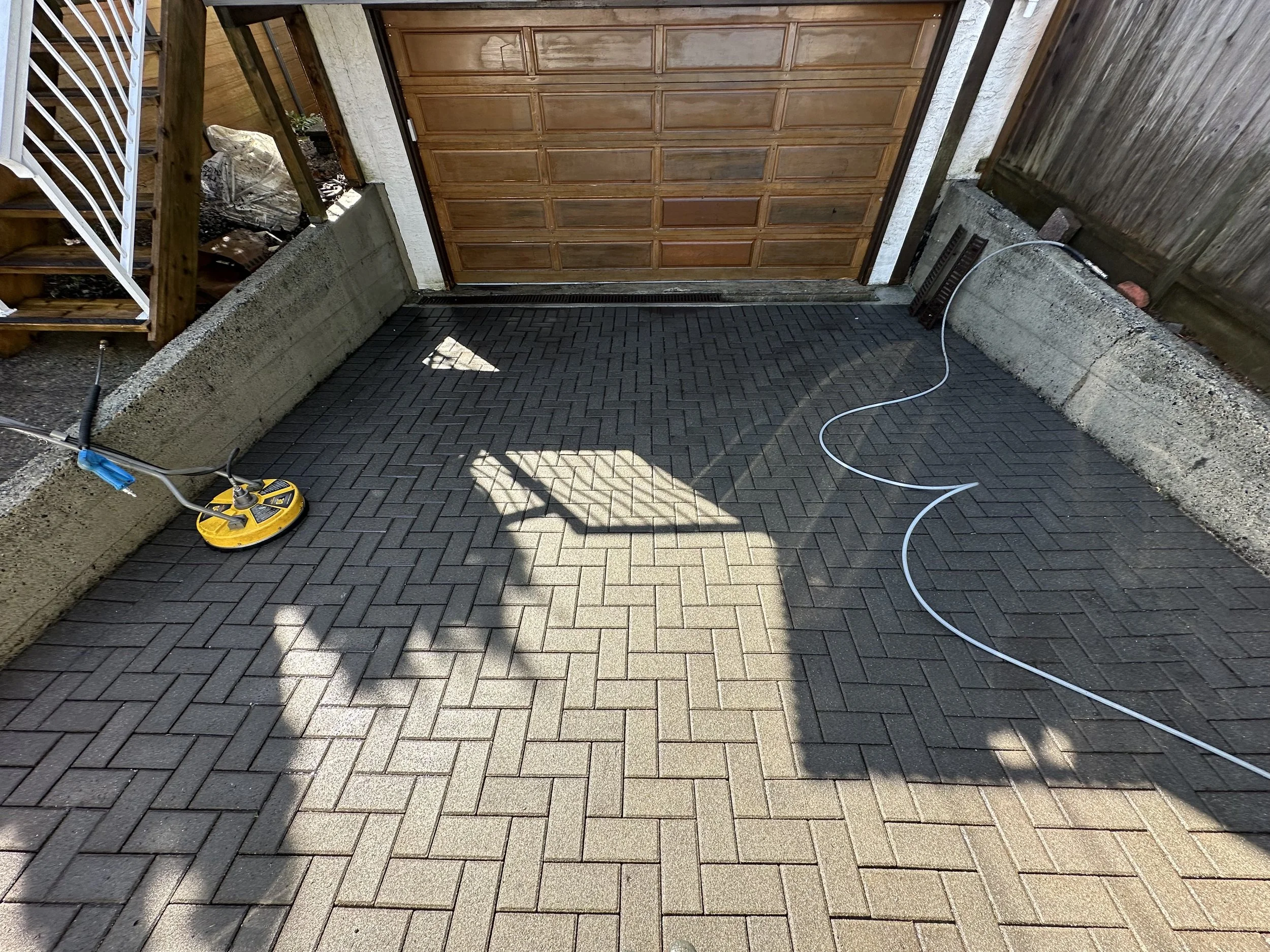 A newly paved brick patio or driveway with dark interlocking bricks. There is a yellow and black vibrating compactor tool on the left side and a coiled white power cord on the right side. The background has a wooden staircase, a closed wooden garage 