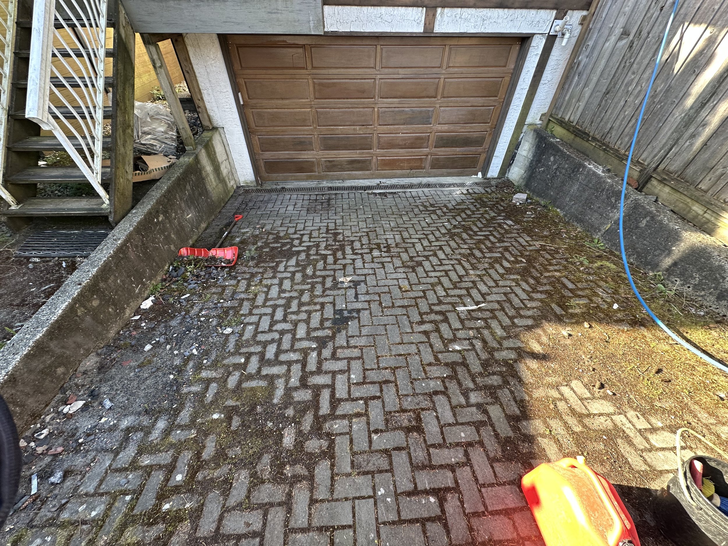 A small paved driveway with a brown garage door in the center, surrounded by a concrete wall on the left and a wooden fence on the right; there is a blue hose on the right side, and a red garden tool handle in the bottom right corner.