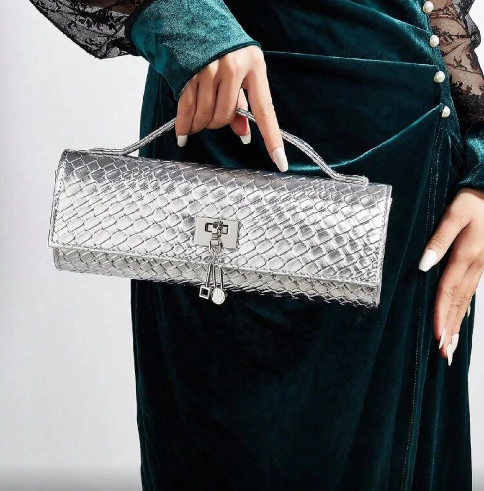 A woman holding a silver, snake-embossed handbag with a metallic clasp and chain handle, dressed in a dark teal velvet dress with black lace sleeves adorned with pearls.