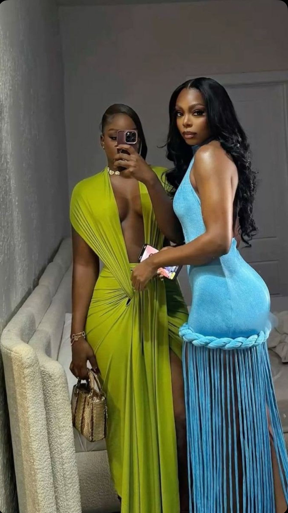 Two women taking mirror selfies, one in a vibrant yellow dress with a deep neckline, and the other in a tight-fitting blue dress with a braided fabric detail at the bottom, in a room with beige walls and a textured armchair.