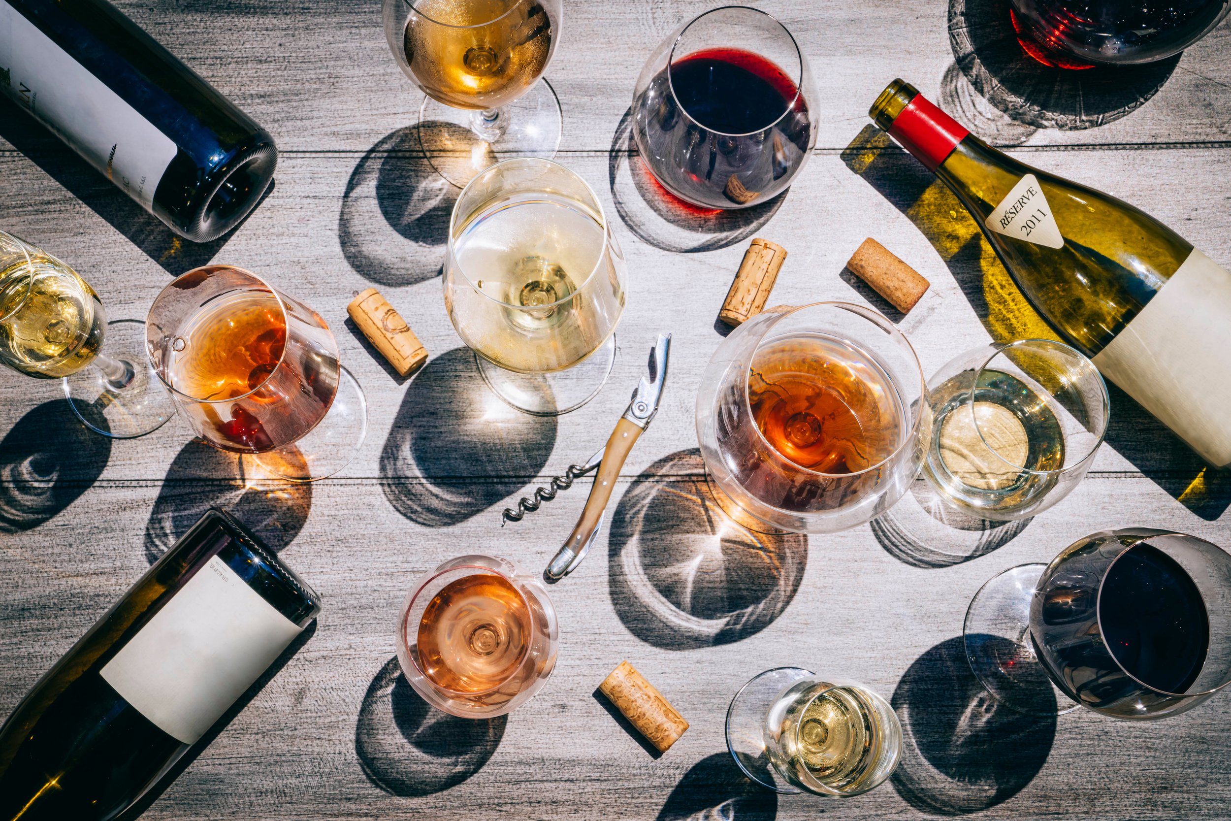 Various wine glasses filled with white, red, and rosé wines on a wooden table, along with wine bottles and corks.