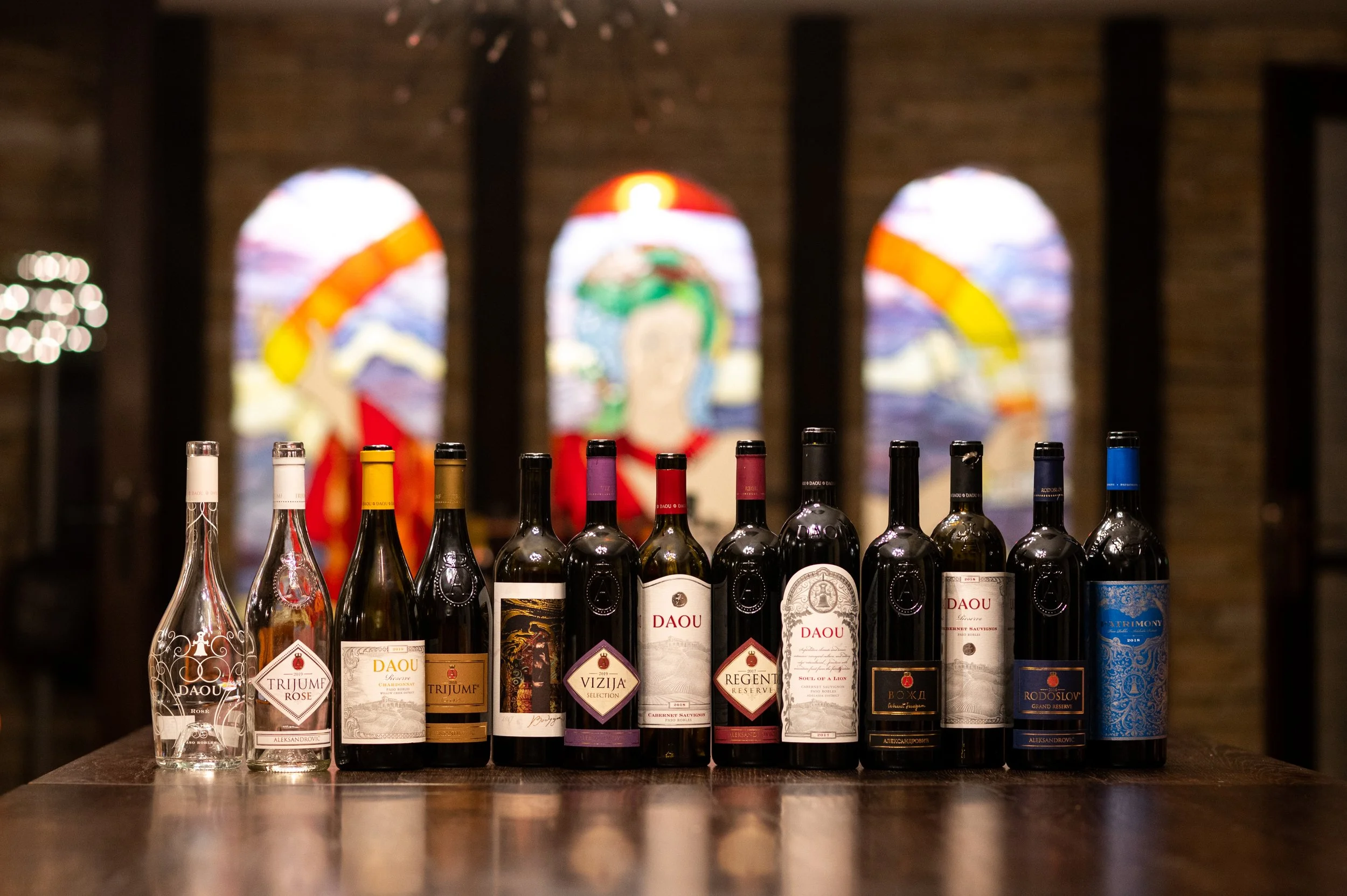 A line-up of various wine bottles with colorful labels on a dark wooden surface, with three arched stained glass windows displaying abstract designs in the background.