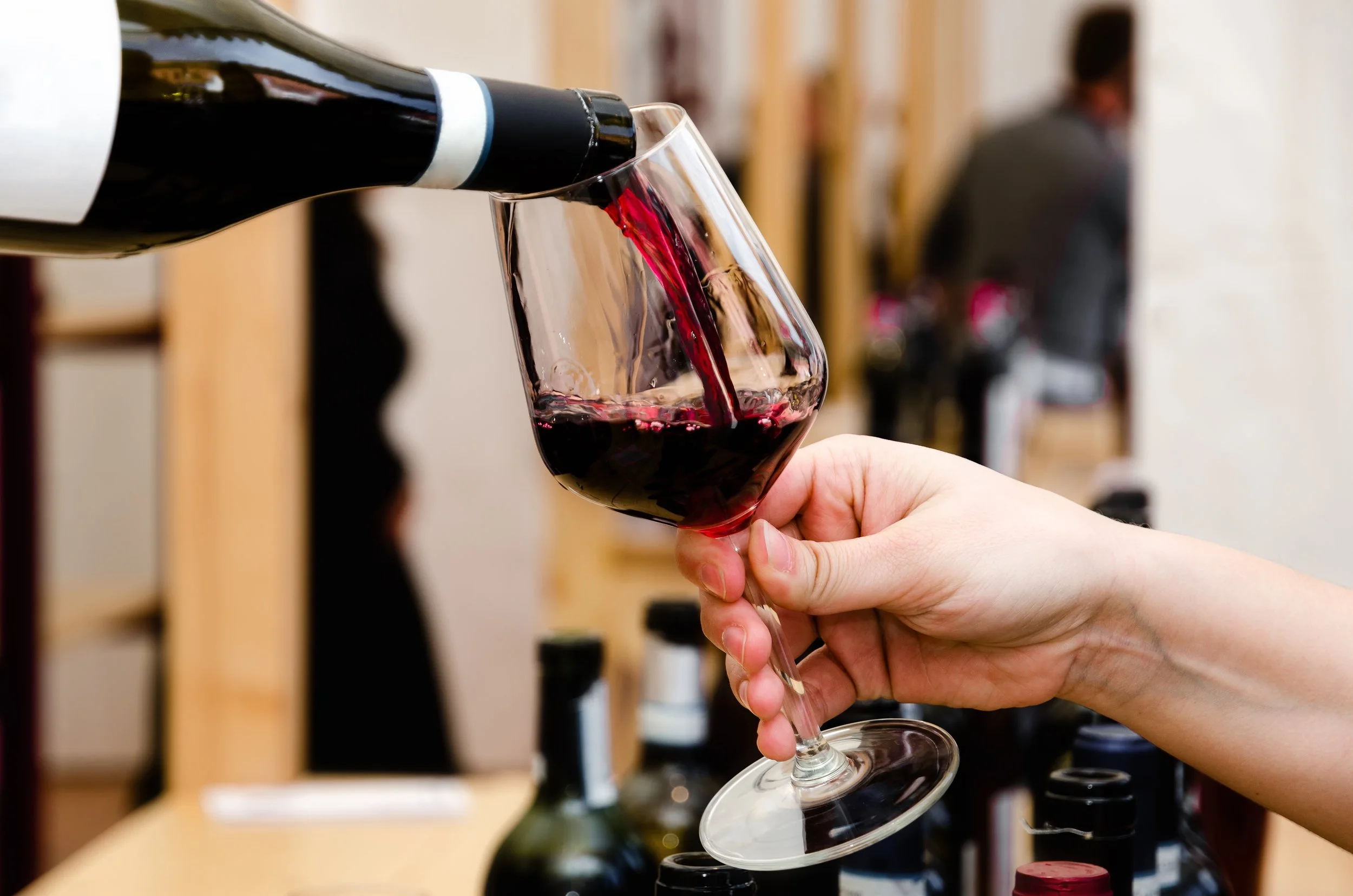 A person pours red wine from a bottle into a wine glass at a wine shop or tasting room.