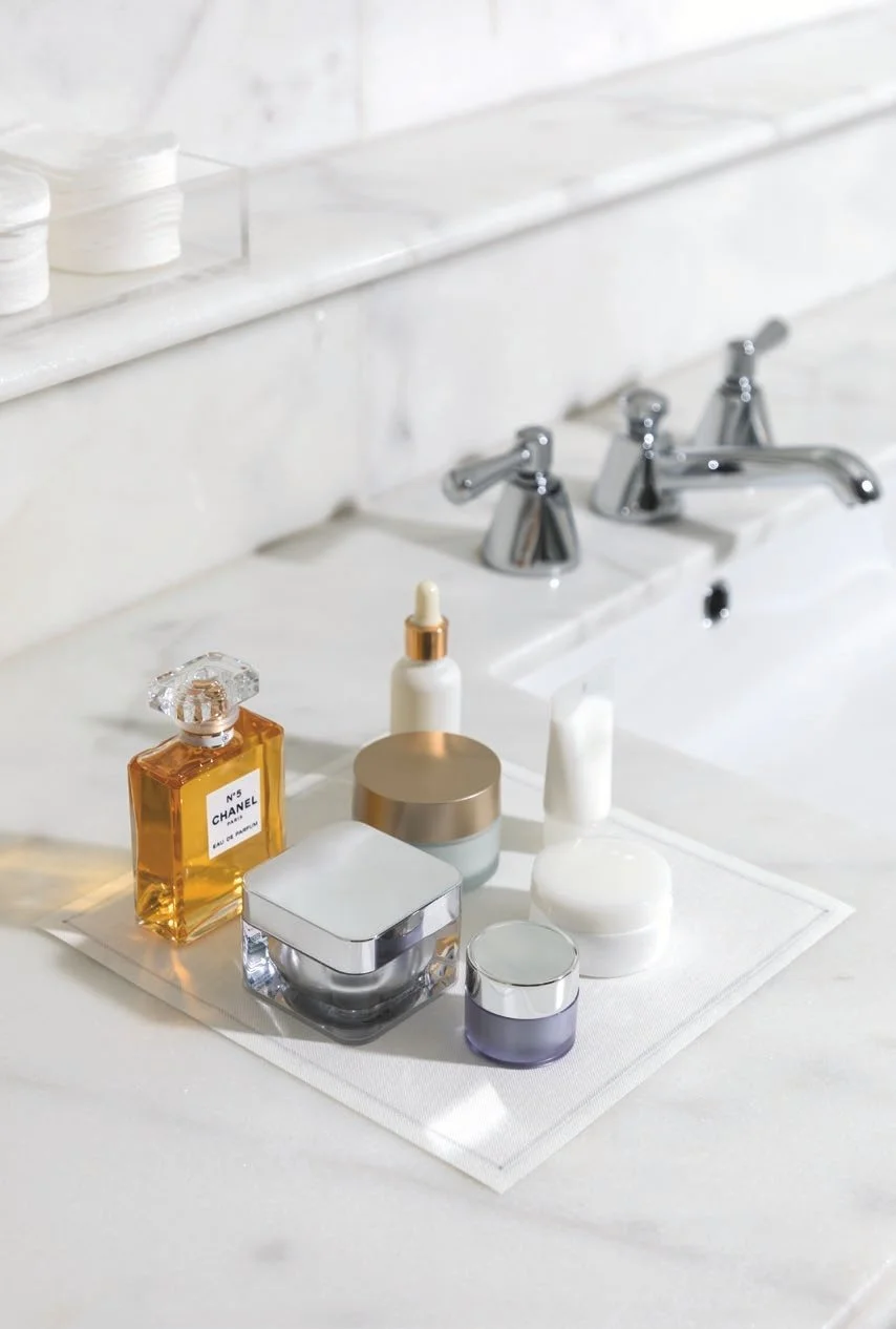 Collection of skincare and perfume bottles on a white mat near a marble bathroom sink with three chrome faucets.