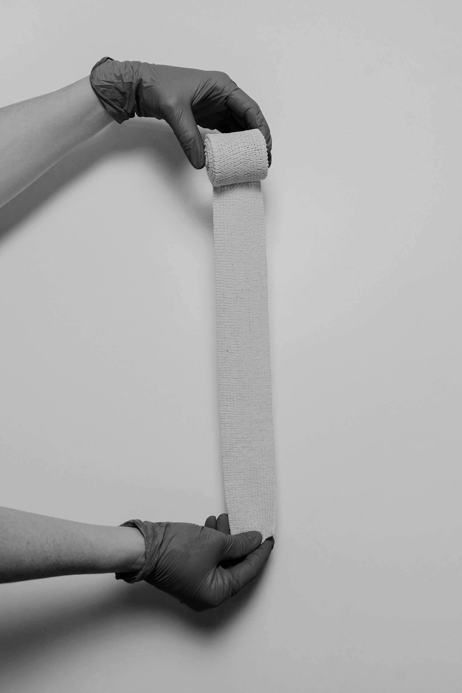 A person wearing black gloves is holding a piece of medical or sports tape, with a roll of tape positioned in the person's hand and the tape hanging down.