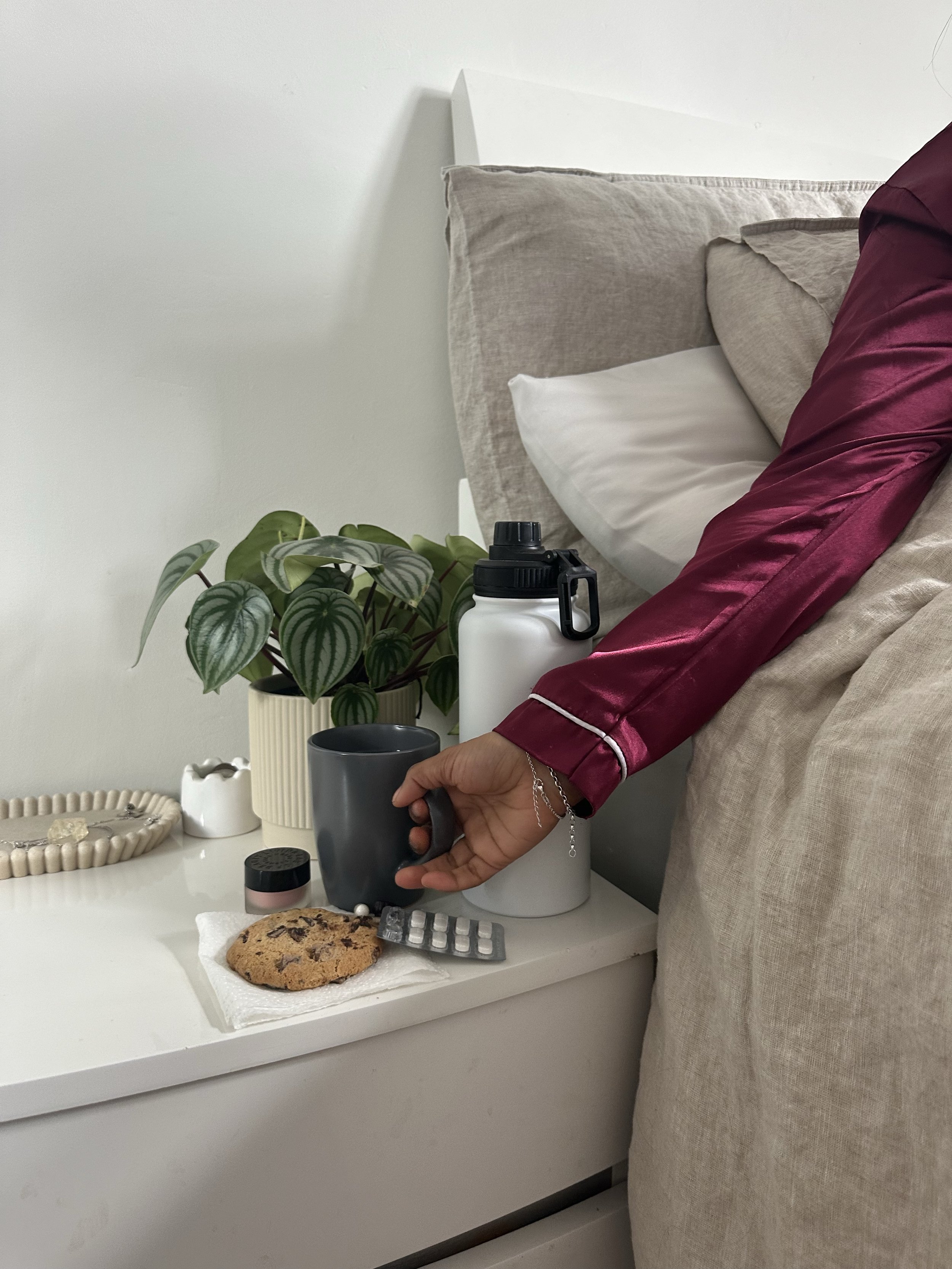 A person wearing a maroon satin robe reaching for a gray mug on a white nightstand, which has a plate with a chocolate chip cookie, a small jar, a small black container, and a box of chocolates. There is a potted plant and decorative items on the nightstand, with a bed and beige pillows in the background.