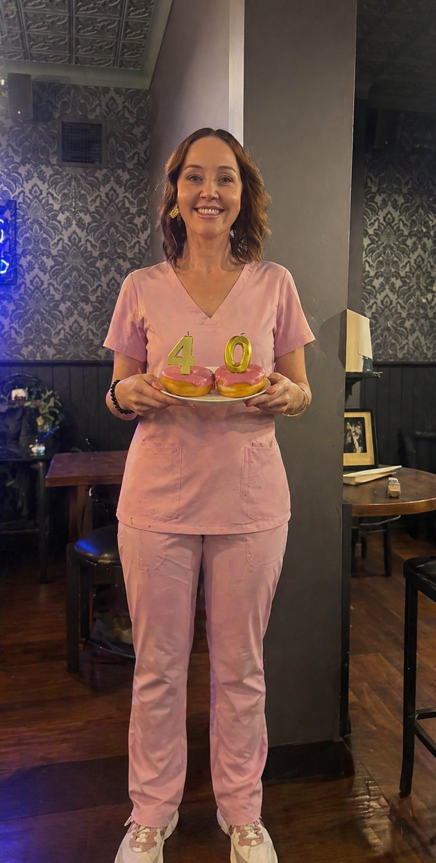 The founder in pink scrubs holding a plate with two donuts and candles shaped as the number 40, celebrating her 40th birthday in a restaurant.