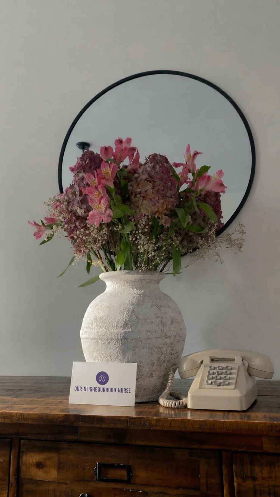 A large white textured vase filled with pink and purple flowers, placed on a wooden surface. A small card reading 'Our Neighbourhood Nurse' is positioned in front of the vase. A beige vintage rotary phone sits beside the vase. A round wall mirror is hung behind the arrangement.