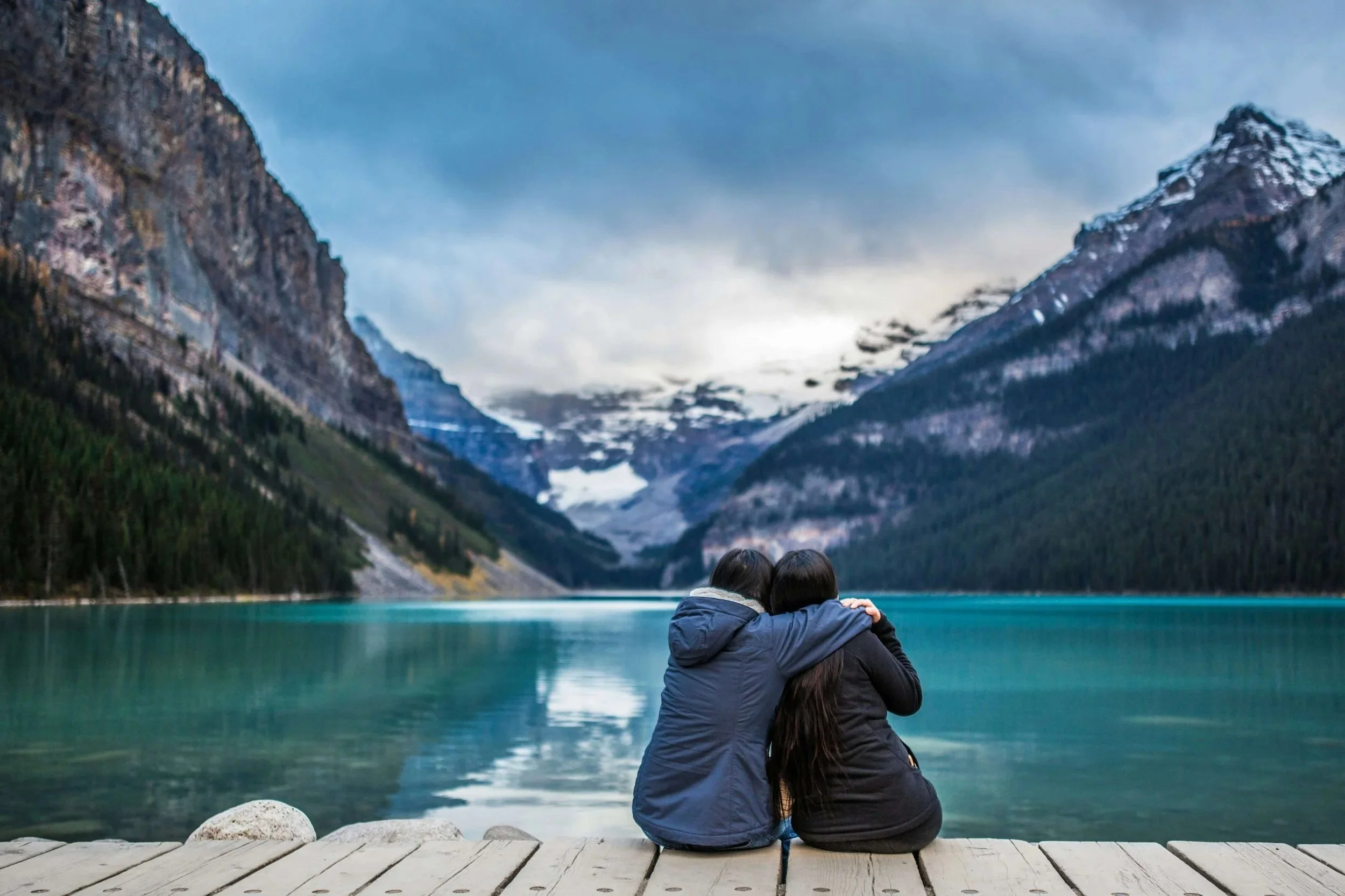 Two people sitting close together on a wooden dock at Lake Louise, turquoise water and snow-capped mountains stretching behind them, quiet and still.