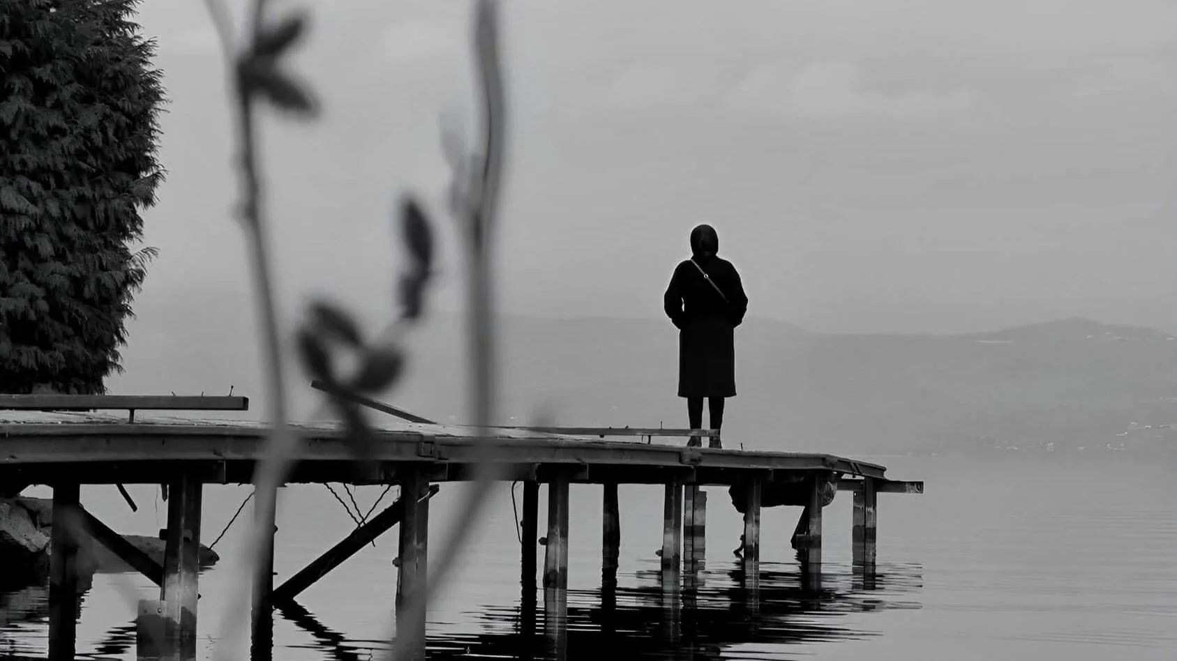 Solitary figure standing at the end of a dock in black and white, grey sky, calm water, trees in foreground