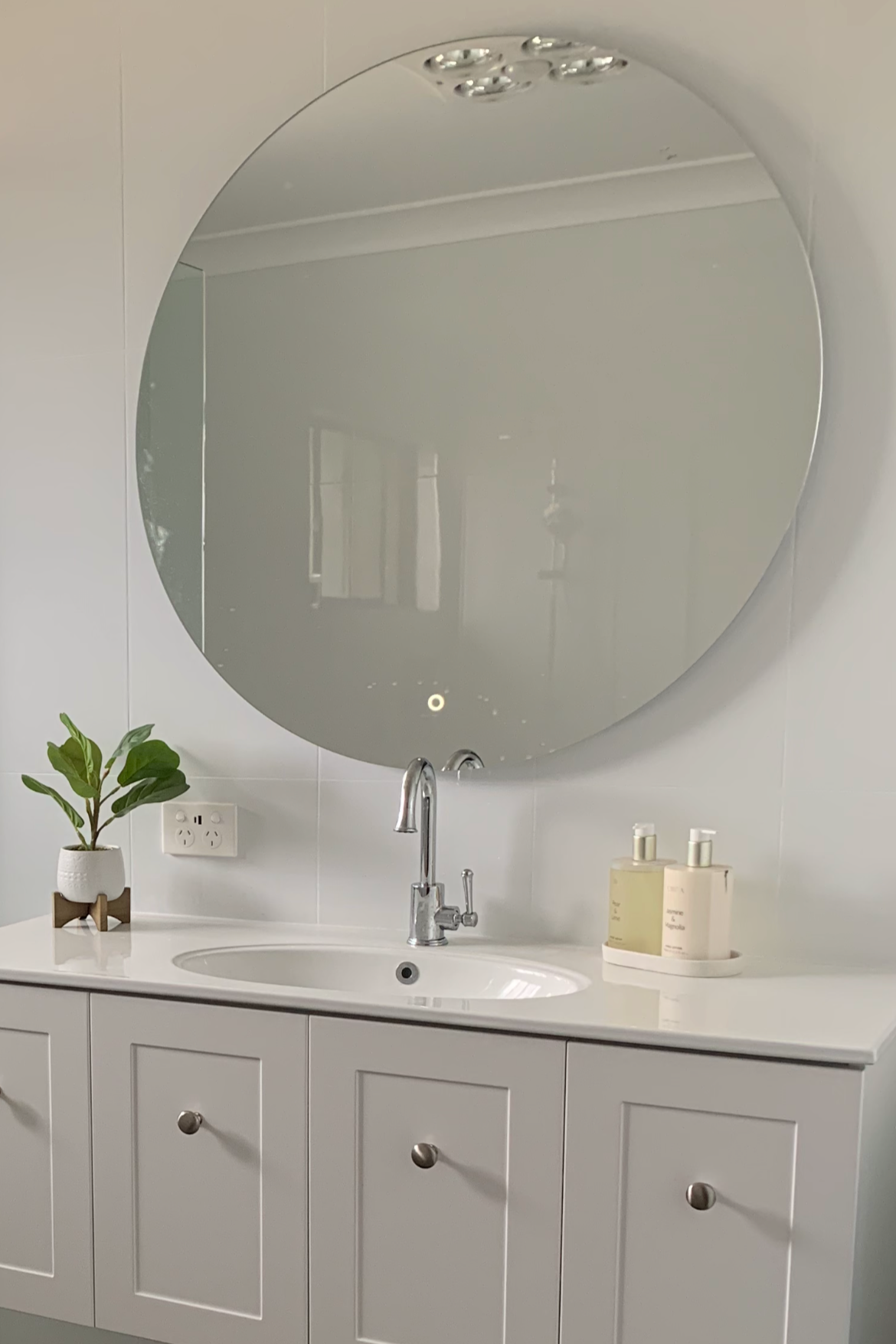 Bathroom vanity with white cabinet, a round mirror, a faucet, a potted plant, and two bottles of lotion or soap on a tray.