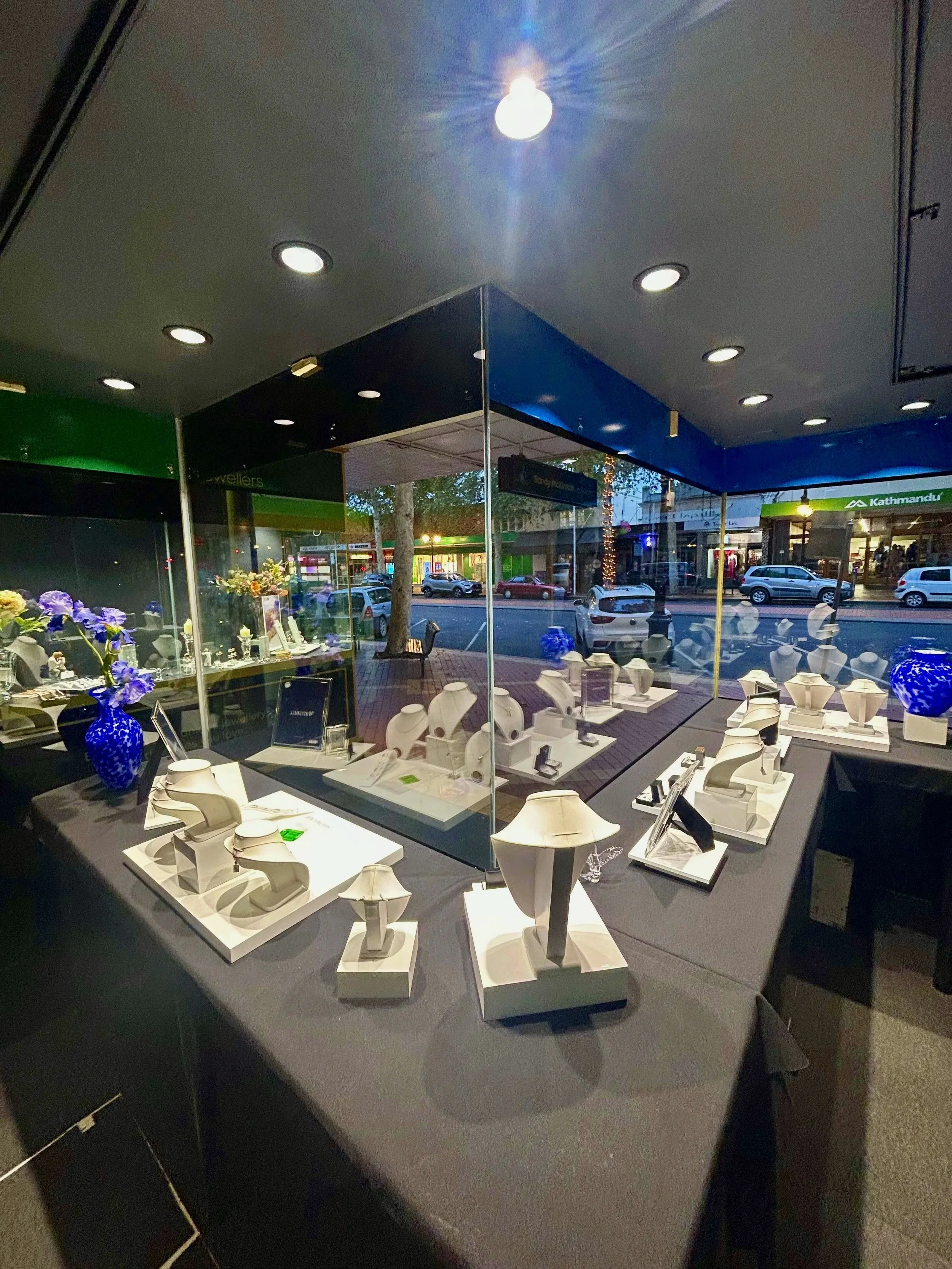 Jewelry display glass case featuring necklaces and rings in a store window, with cars and retail stores visible outside.