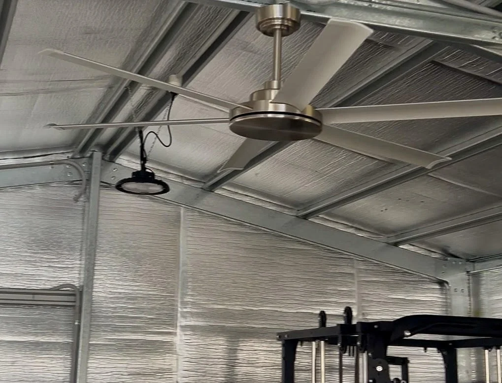 Interior of a metal garage or warehouse with a ceiling fan, a hanging light fixture, and exercise equipment.