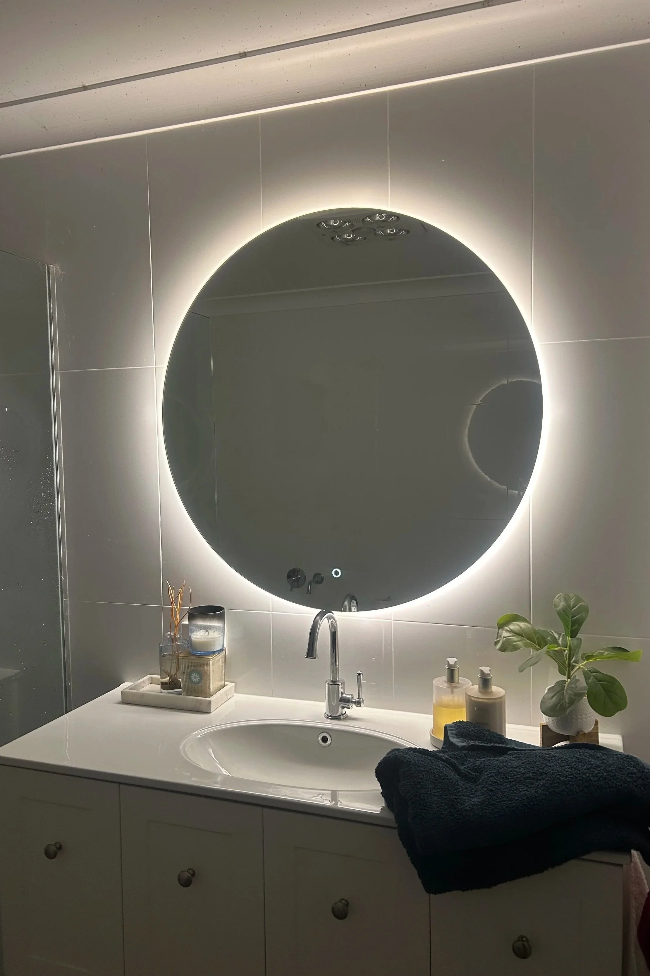 Bathroom vanity area with a round backlit mirror, a white countertop with a modern sink, faucet, and soap dispensers, a small tray with decorative items, a potted plant, some towels, and various toiletries.