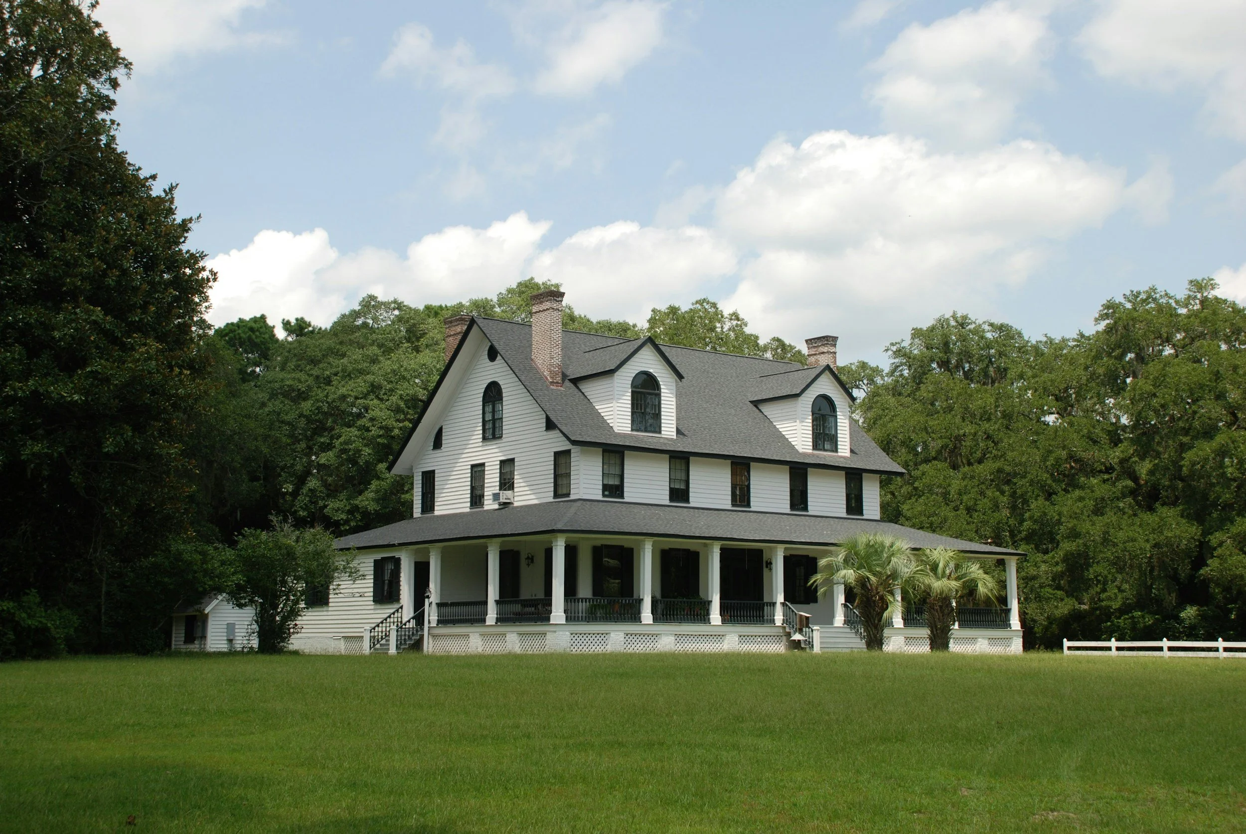 A large white house with black window frames and a dark roof, surrounded by trees and a green lawn under a partly cloudy sky.