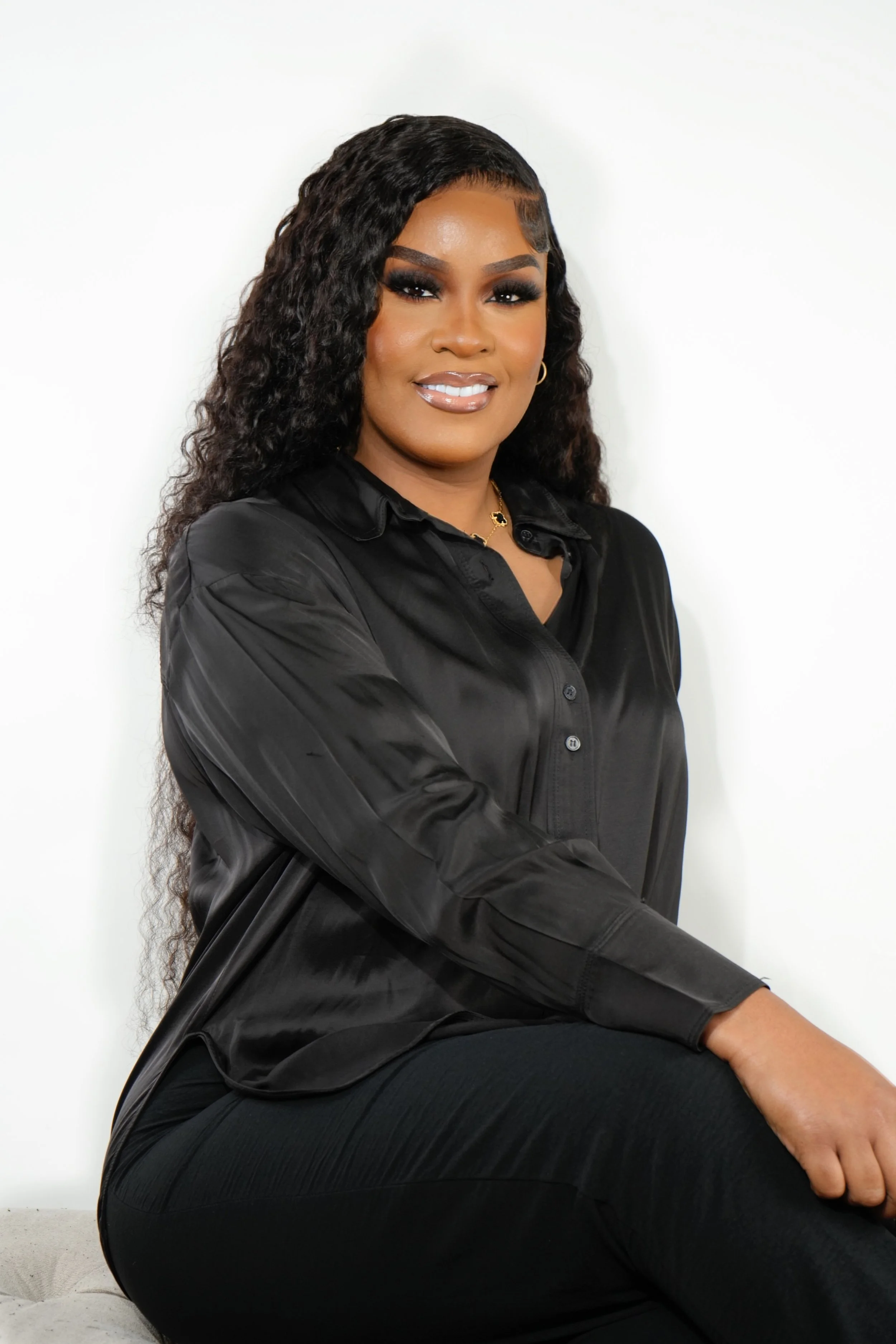 A woman with long curly black hair, wearing a black silky blouse and black pants, smiling while sitting against a plain white background.