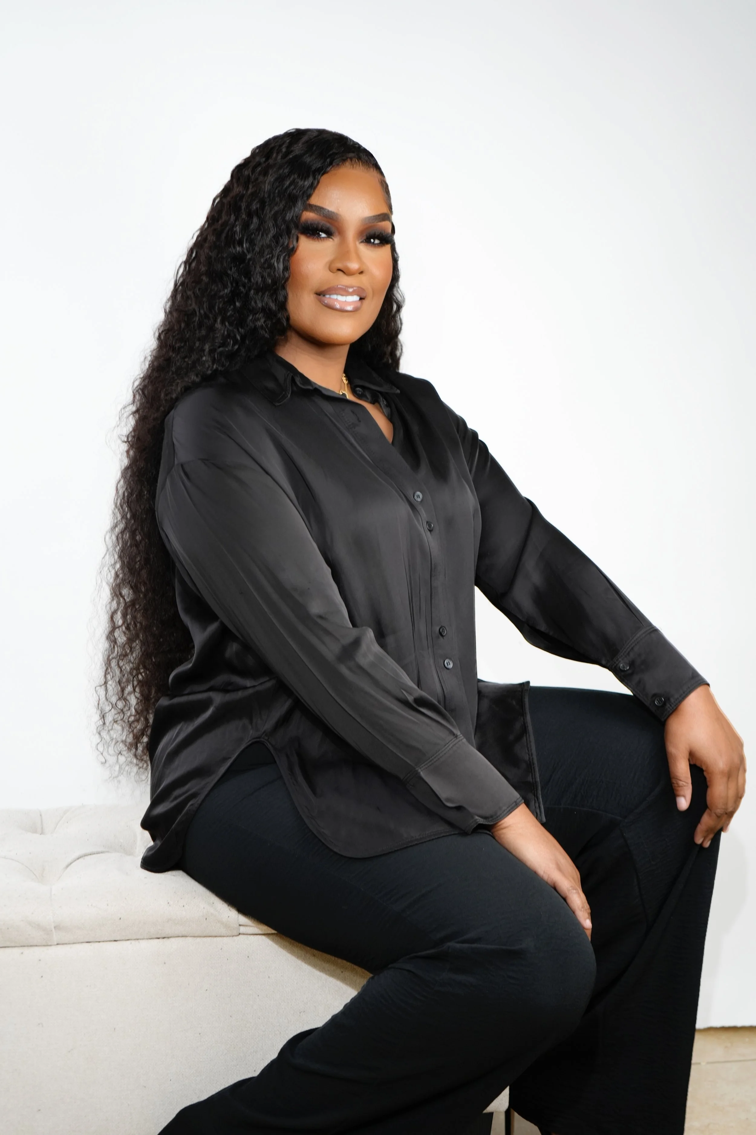 A woman with long curly hair and makeup, wearing a black satin button-up shirt and black pants, sitting on a white bench against a plain white background.