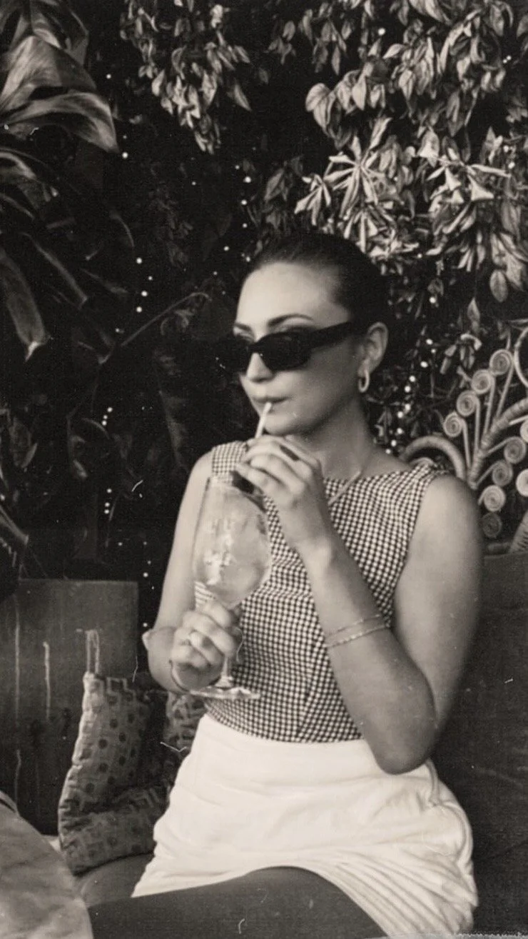 A woman with dark hair pulled back, wearing sunglasses, a checked sleeveless top, and a white skirt, drinks from a large glass with a straw, holding it with one hand.