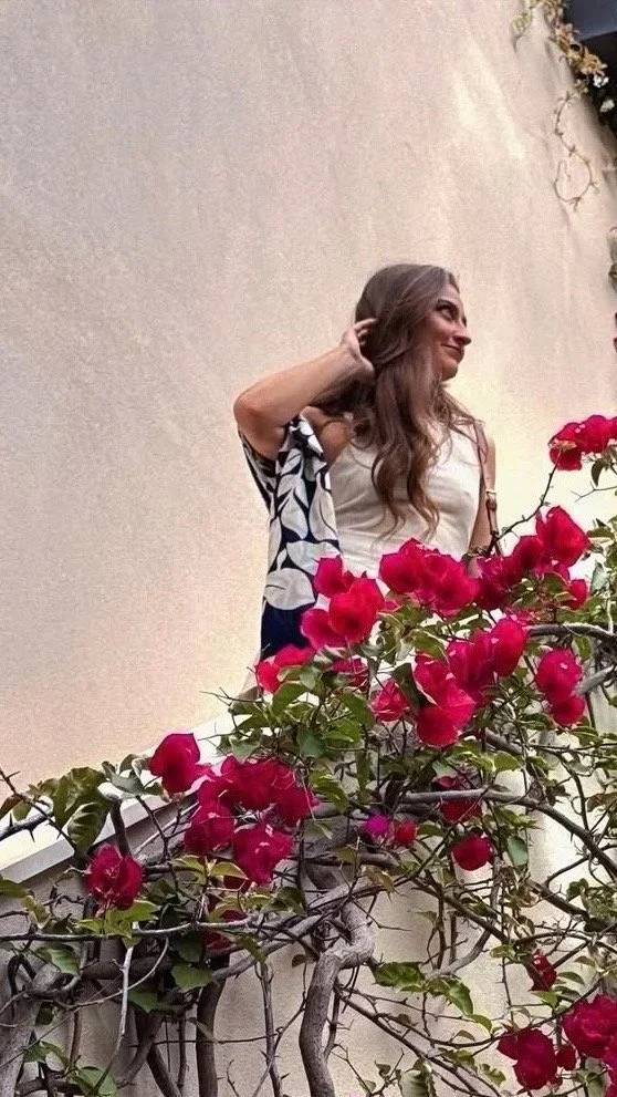 A woman standing outdoors next to a wall covered with pink bougainvillea flowers, smiling and looking to the right.