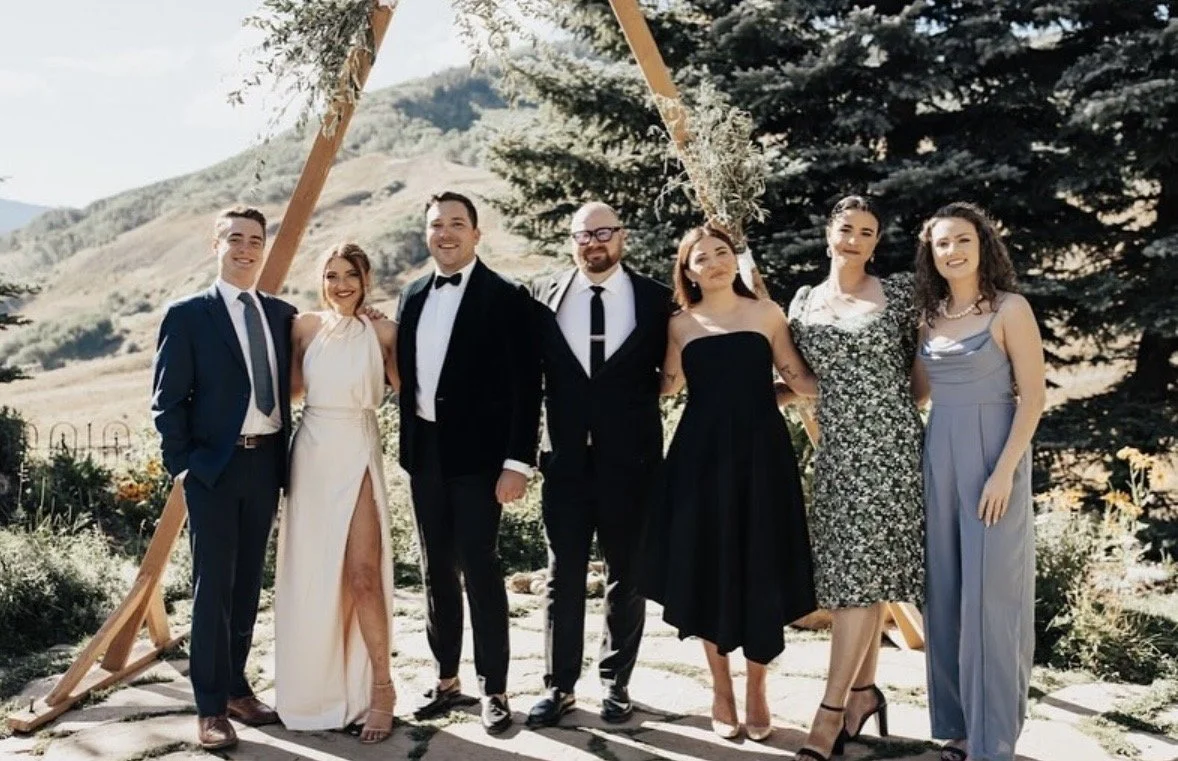 Group of seven people dressed in formal attire standing outdoors near a wooden wedding arch with mountains and trees in the background.