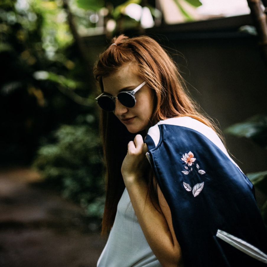 A young woman with red hair wearing sunglasses and a white top, holding a navy blue jacket with floral embroidery over her shoulder, standing outdoors surrounded by greenery.