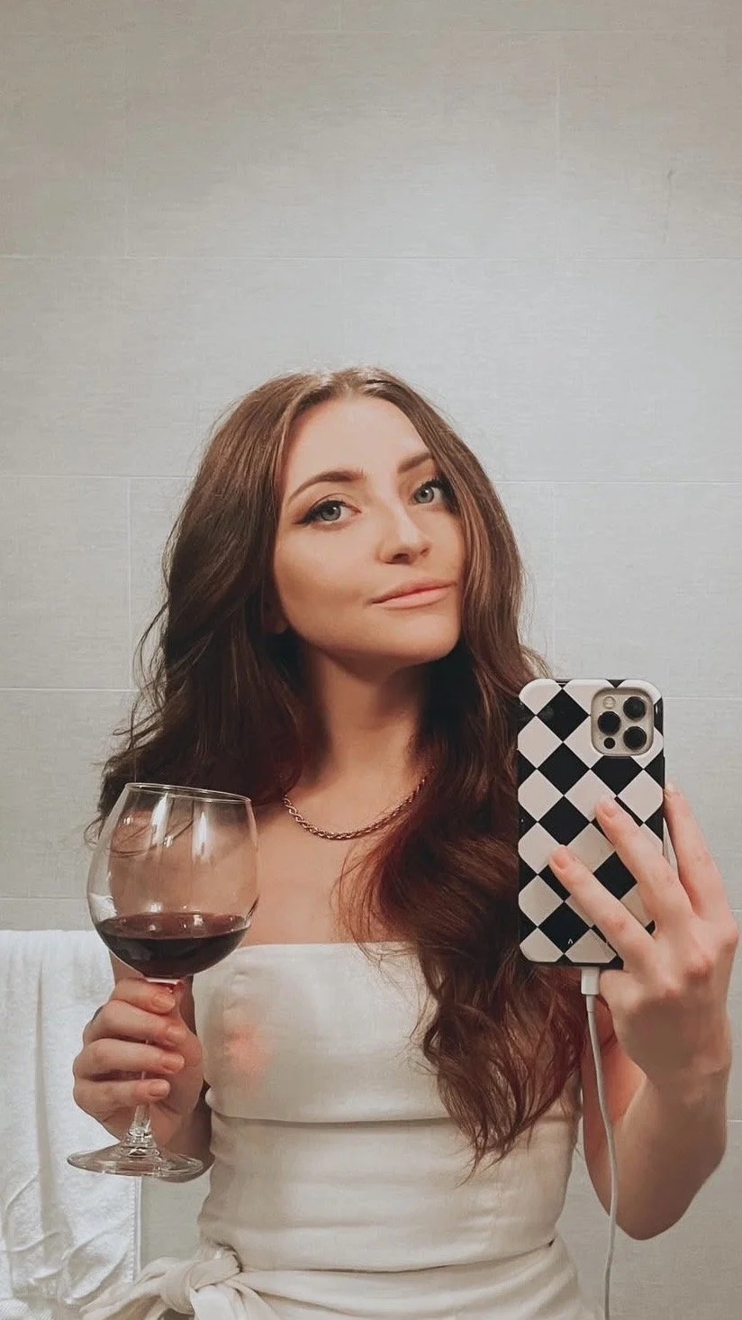 A woman with long red hair taking a selfie in a bathroom mirror, holding a glass of red wine in her left hand, wearing a white dress and a gold chain necklace.