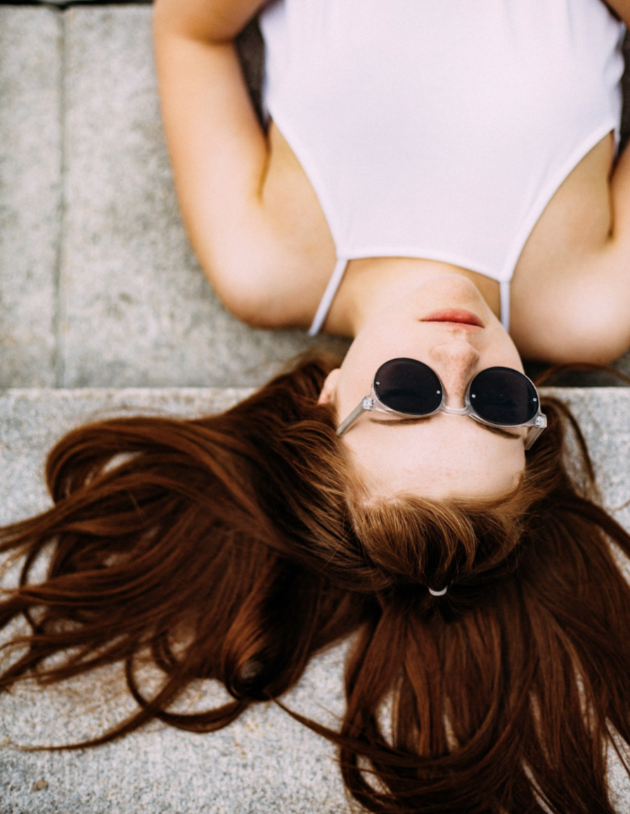 Young woman with long red hair wearing sunglasses and a white sleeveless top, lying on her back on a concrete surface.