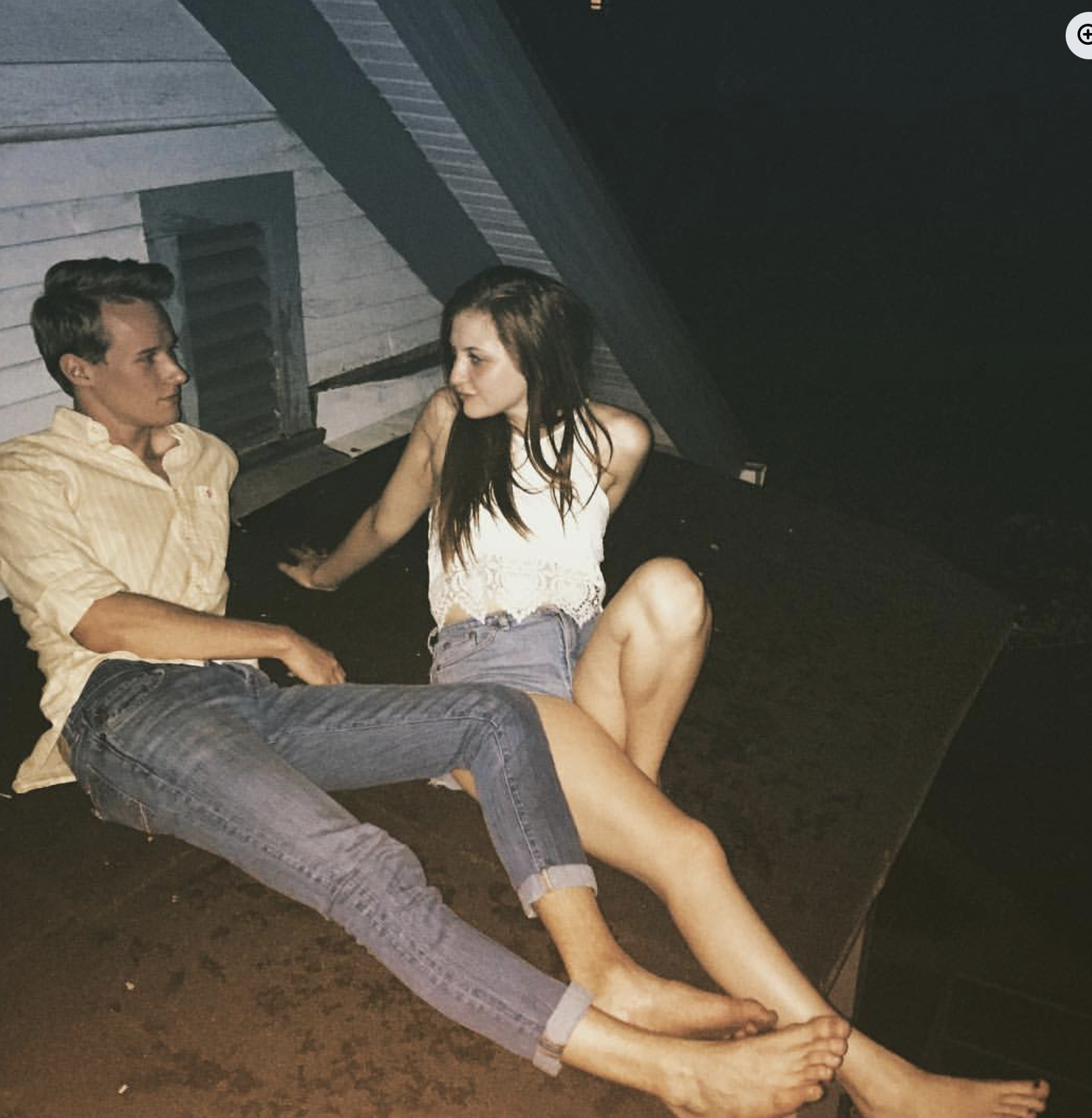 Two young adults, a man and a woman, sitting close together outdoors near a wooden building with painted siding at night. They are casually dressed, and the woman appears to be leaning on the man's shoulder.