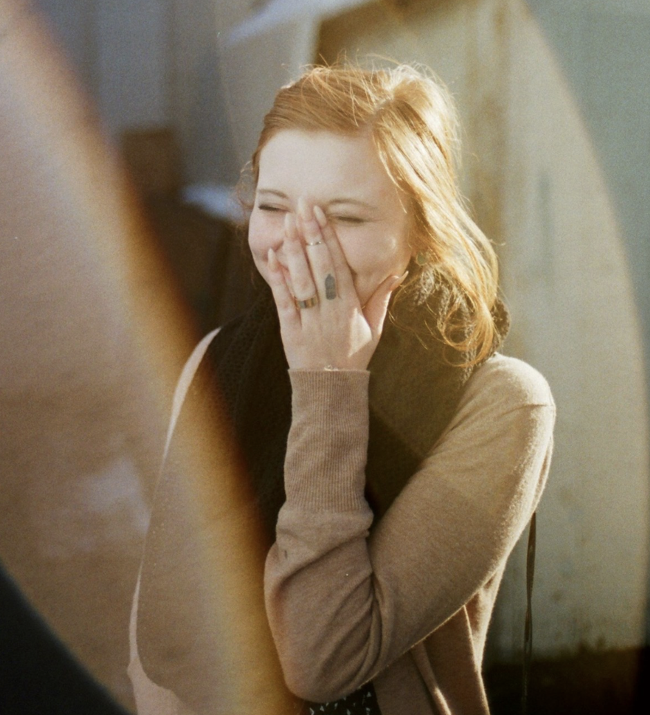 A woman with red hair laughing and covering her mouth with her hand, wearing a beige sweater and rings, in bright sunlight.