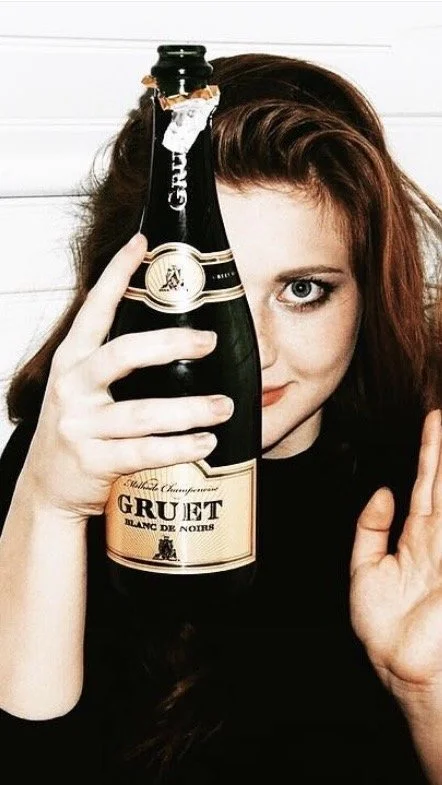 A woman with long brown hair holding a champagne bottle in front of her face, partially covering it. She is smiling and has her right hand raised with fingers near her face, standing in front of a white wooden background.