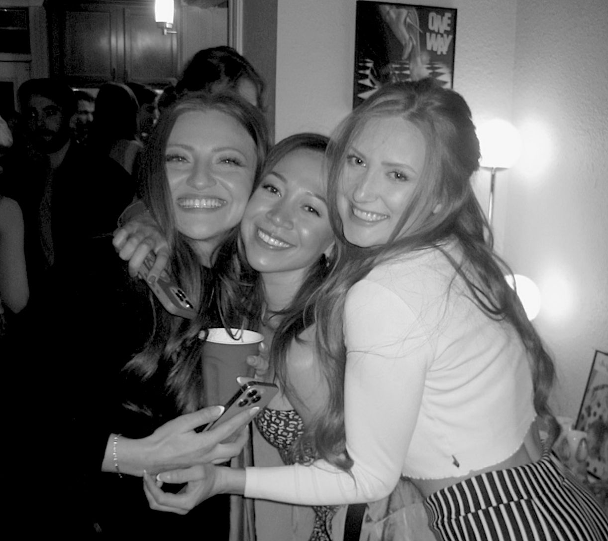 Three young women happily hugging and smiling at a social gathering, with a group of people in the background and a framed poster on the wall.