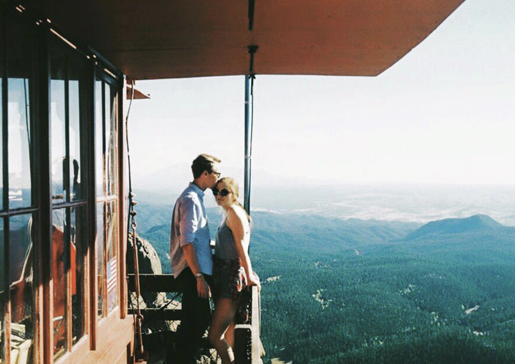 A couple standing on a mountain viewing platform, overlooking lush green mountains and valleys, with clear blue sky in the background.